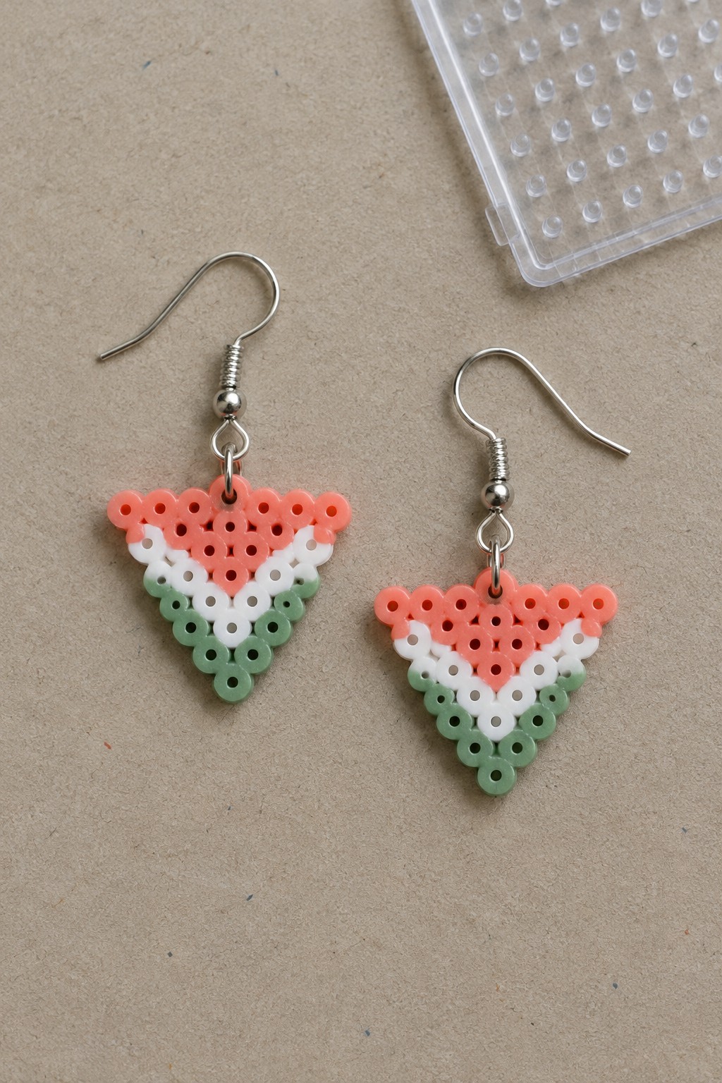 Two perler bead watermelon slice earrings with silver hooks on beige surface.