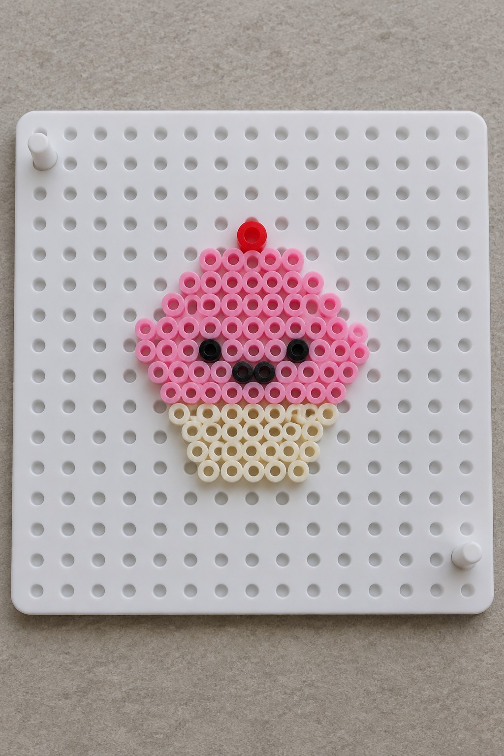 Beaded pink cupcake with face, red cherry, and beige base on white pegboard