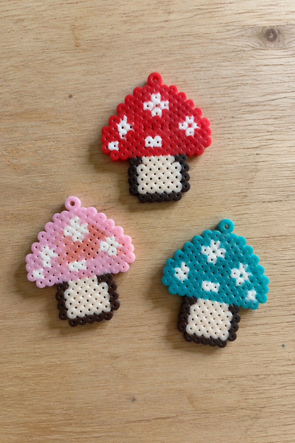 Three perler bead mushroom charms in red, pink, and teal on wood