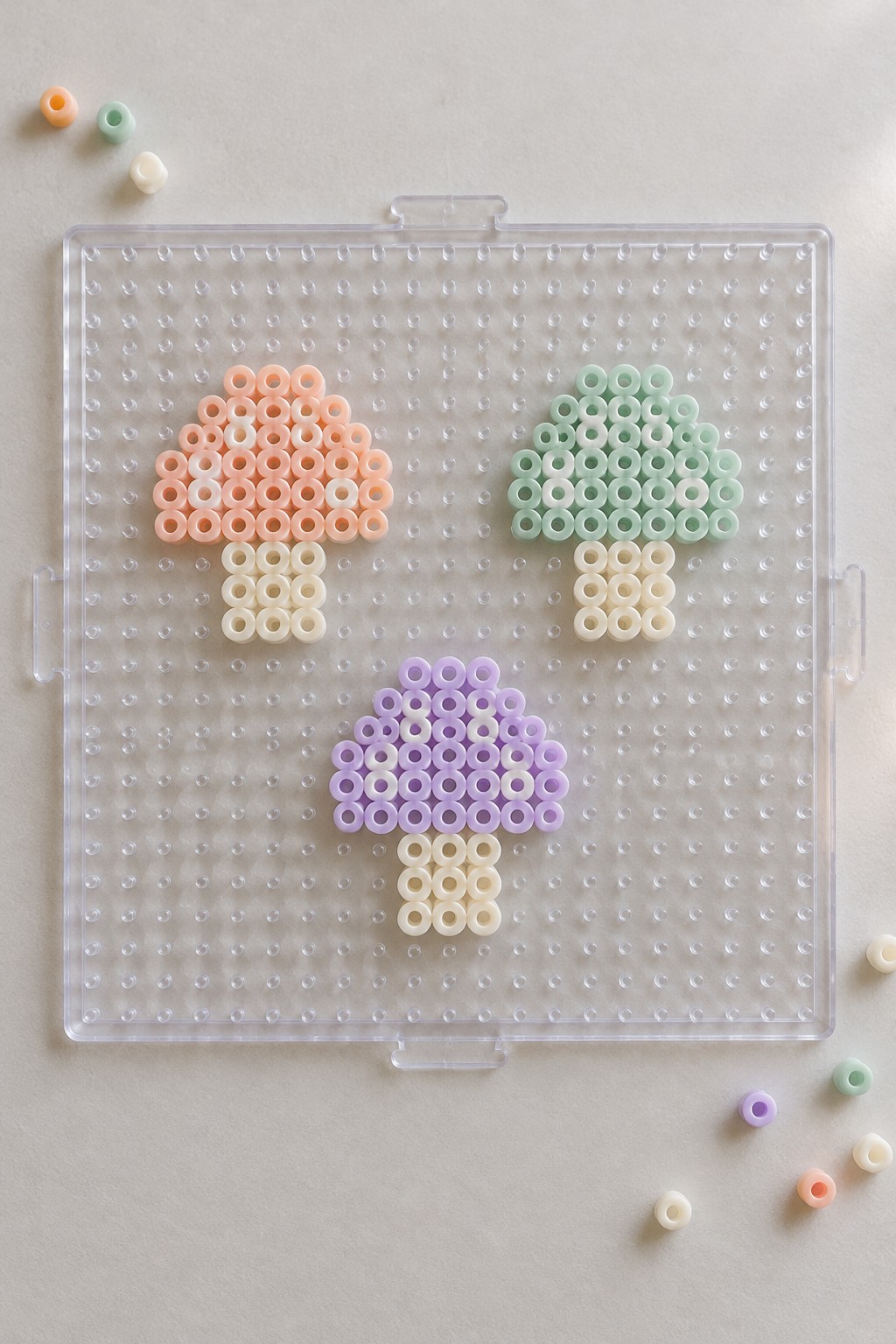 Three pastel perler bead mushrooms on a clear pegboard with loose beads