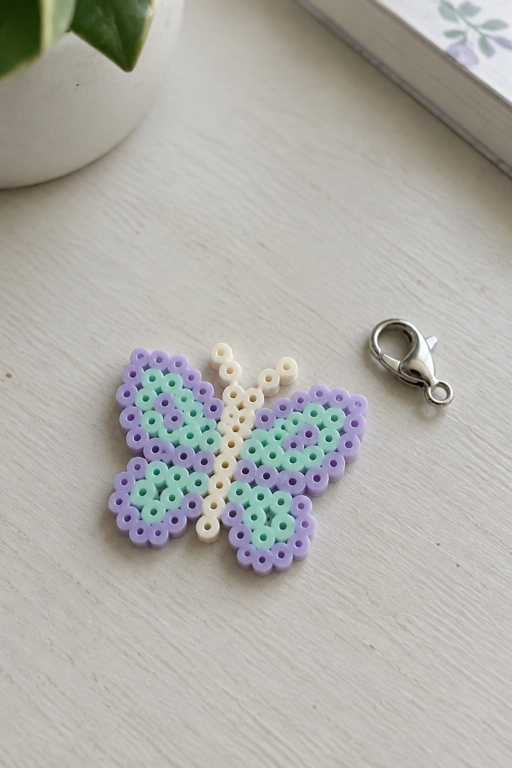 Pastel perler bead butterfly in purple and mint green beside metal clasp on wood