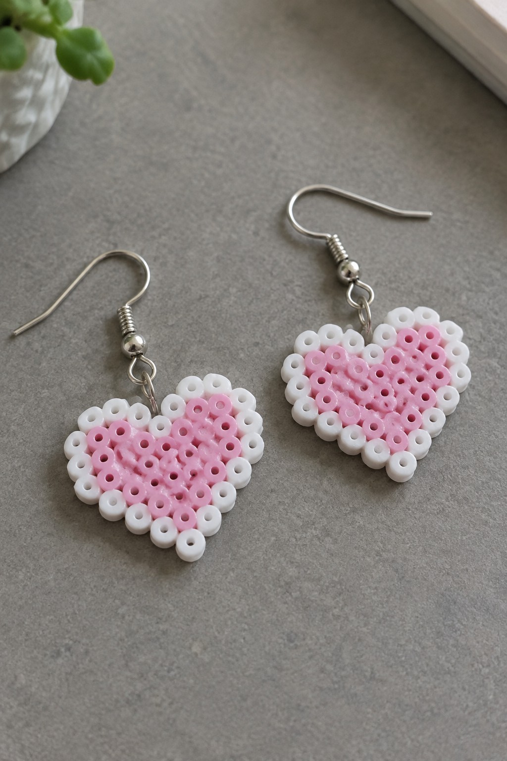 Two small heart-shaped Perler bead earrings in pink and white attached to silver hooks.