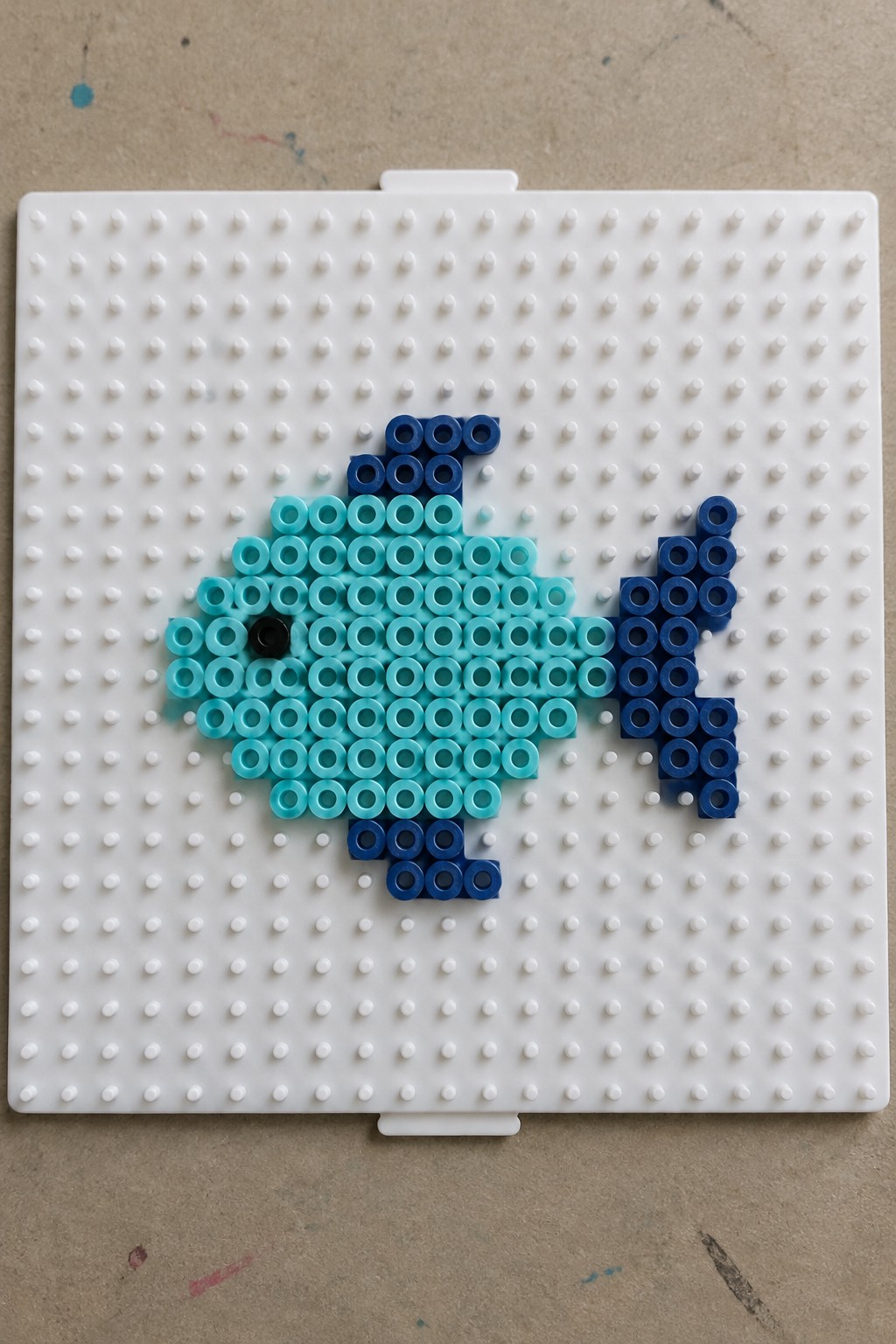 A fish pattern made with light blue and dark blue Perler beads on a white pegboard, with a black bead for the eye.