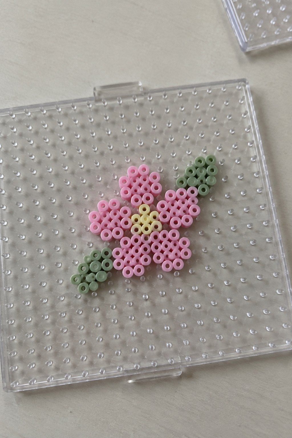 Pink perler bead flower with yellow center and green leaves on clear pegboard.