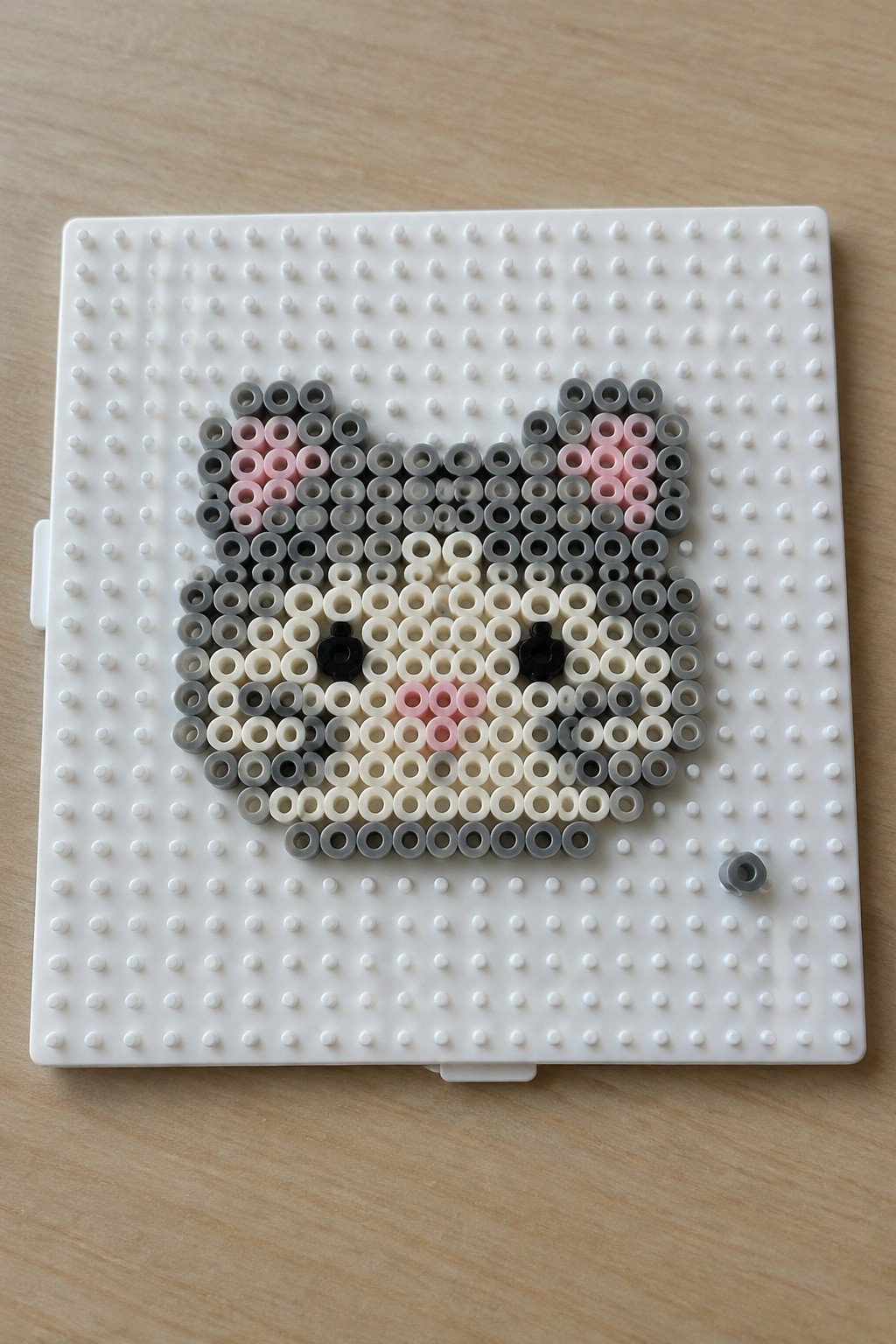 Perler beads forming gray pink cat face on white pegboard with loose bead