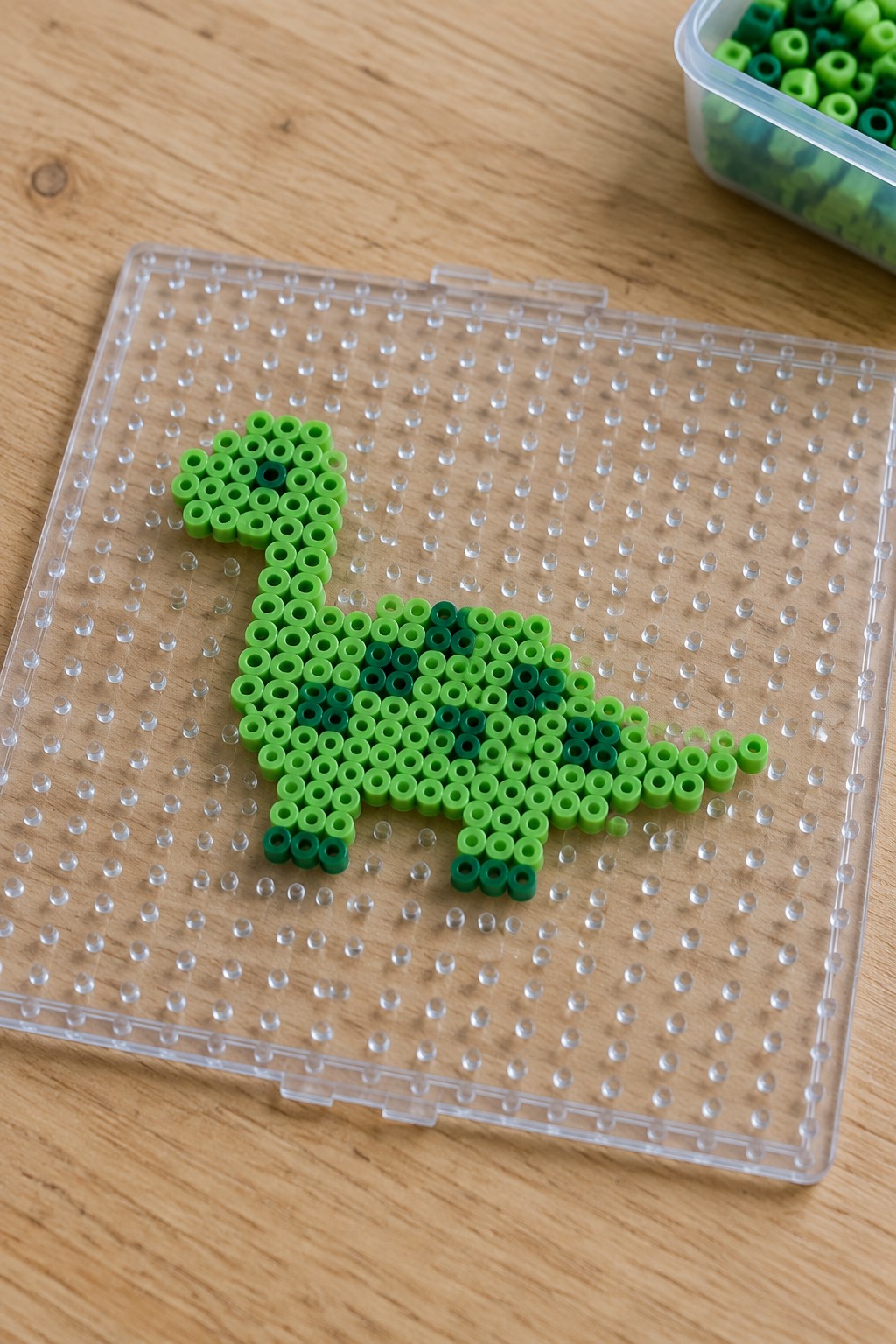 Green perler bead dinosaur on clear pegboard beside bead container on wood
