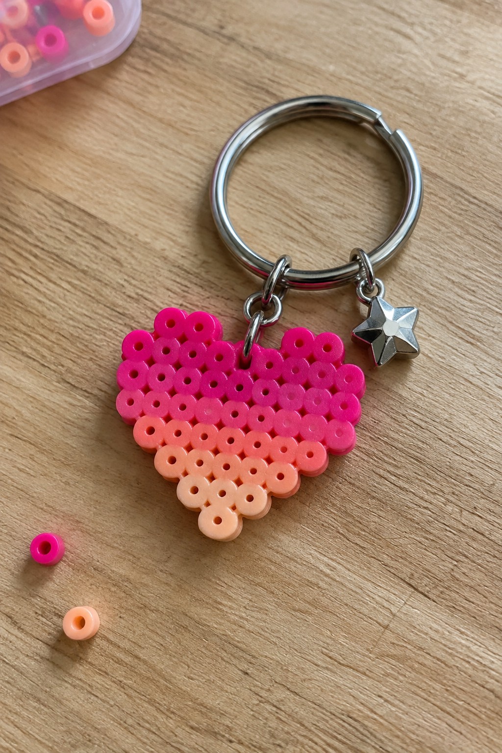 Pink-orange gradient perler bead heart keychain with silver star charm on wood.