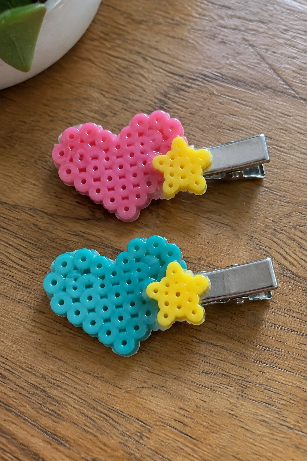 Two fused Perler bead hair clips showing a pink heart with yellow star and a cyan heart with yellow star attached to metal clips.