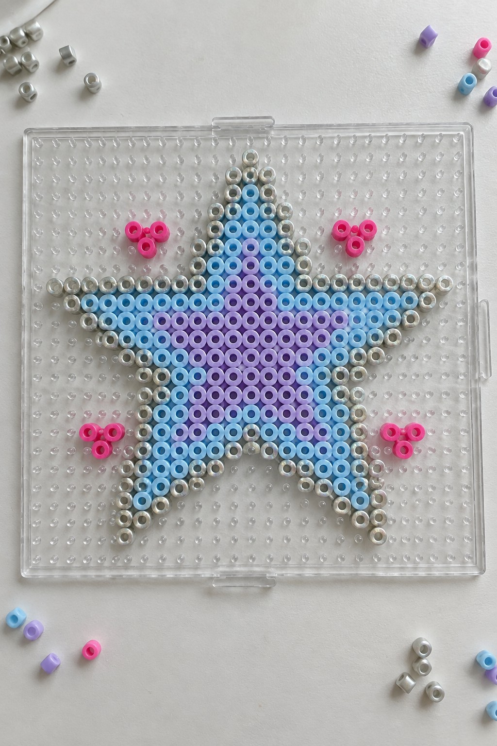 Star-shaped Perler bead pattern in blue, purple, and silver on clear pegboard with pink accents