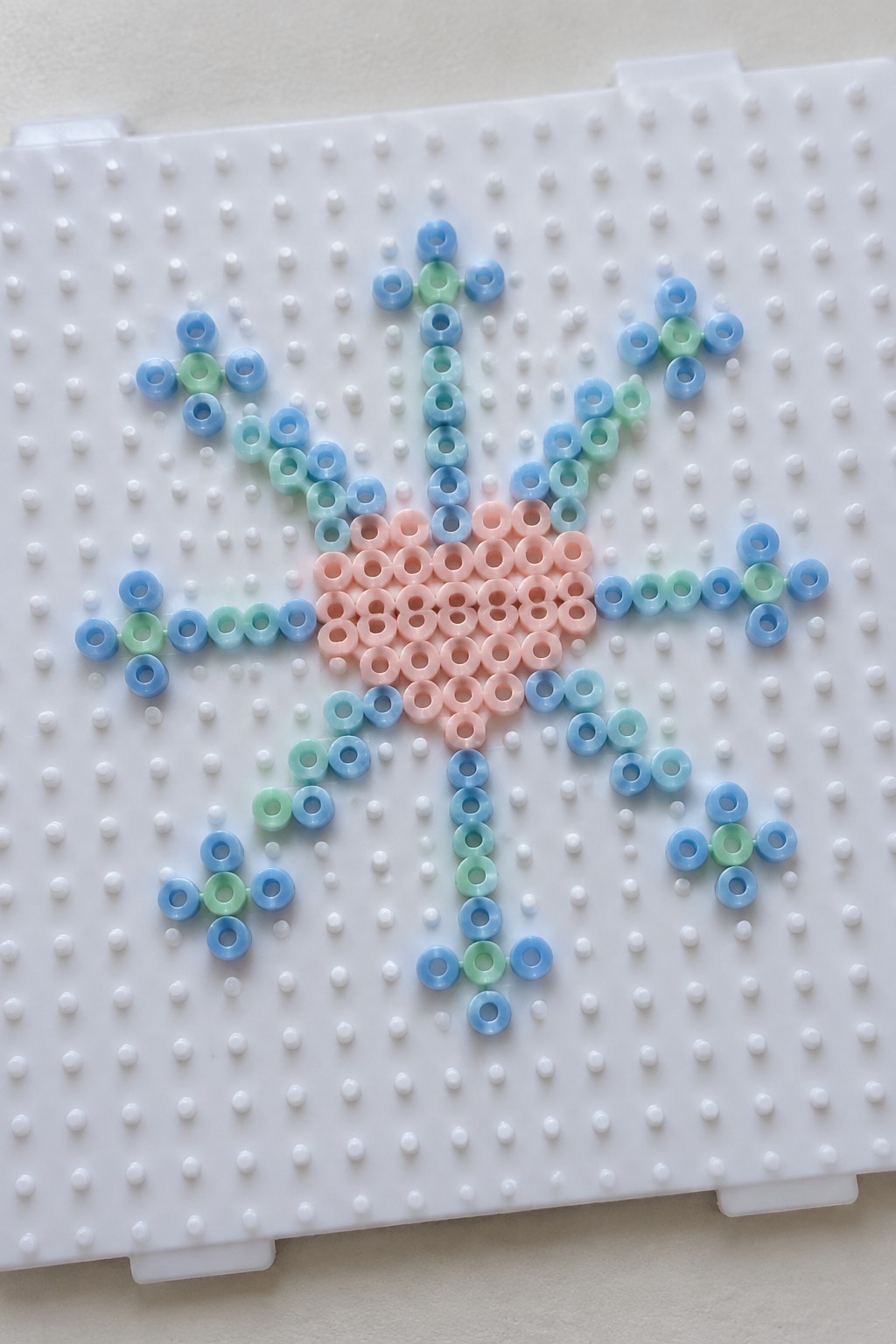 Pastel blue, green, and pink perler beads forming a flower on white pegboard.