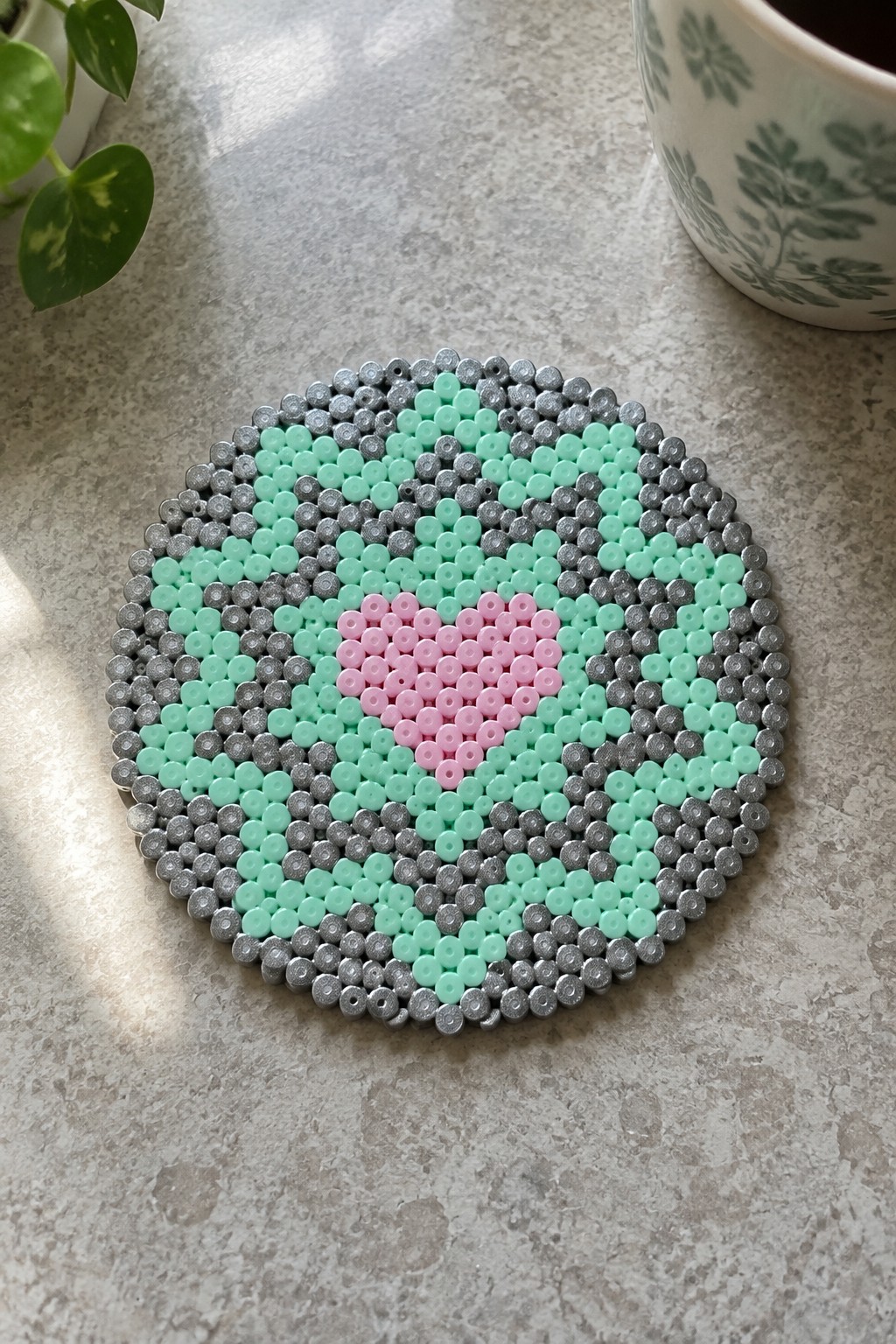 Round perler bead coaster with pink heart center and mint green gray swirls on countertop