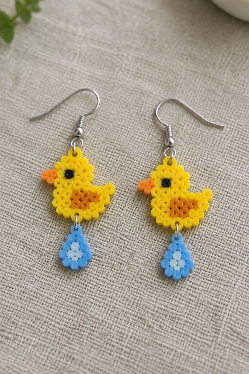 Two yellow Perler bead duck earrings with orange beaks, black eyes, and blue hanging drops on a fabric surface.