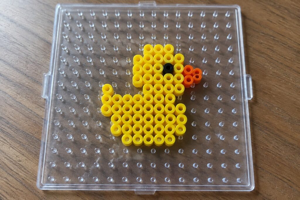 Yellow duck made of beads with black eye and orange beak on clear pegboard.