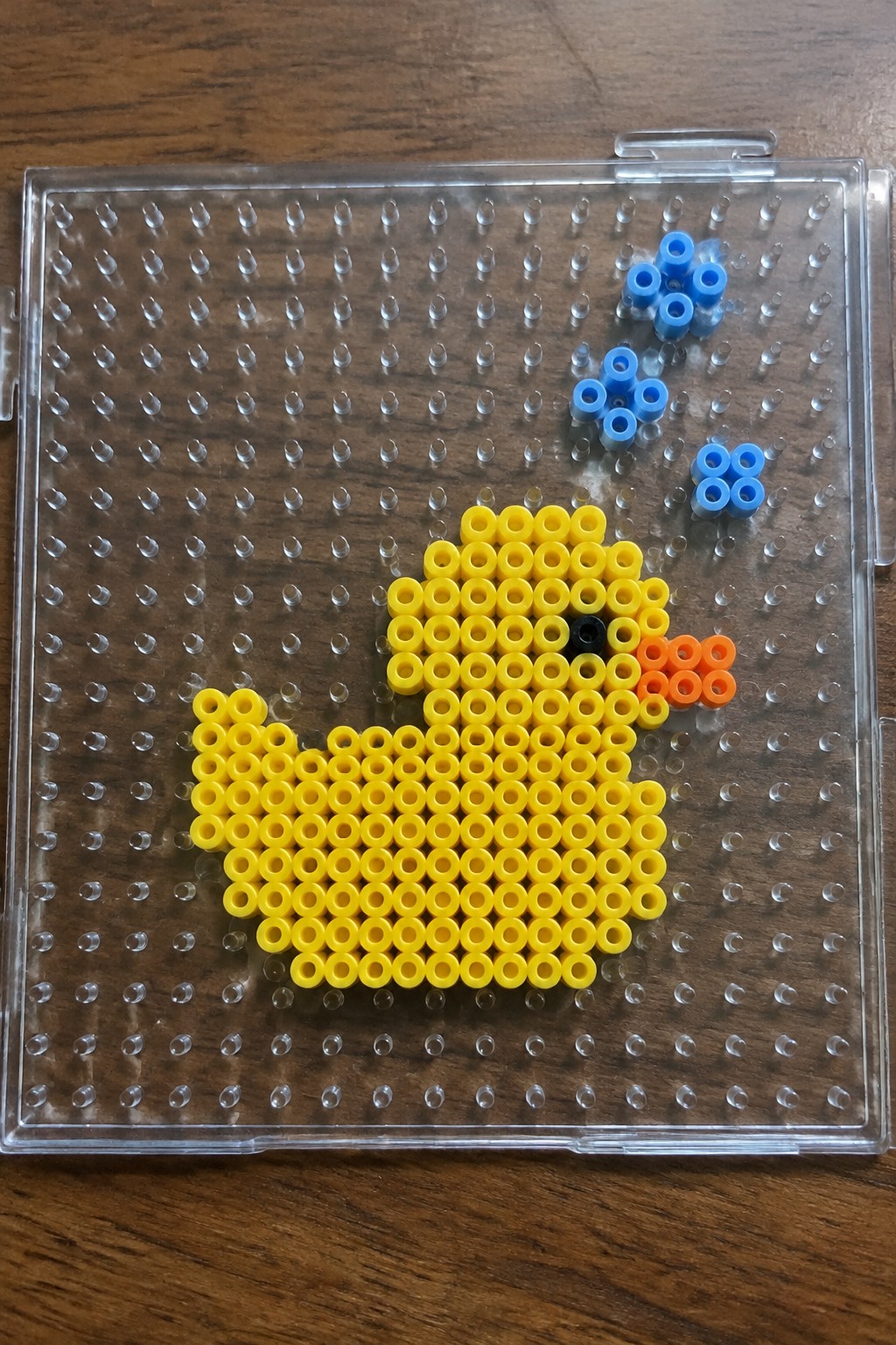 Yellow duck Perler bead pattern on a clear pegboard with black eye and orange beak.