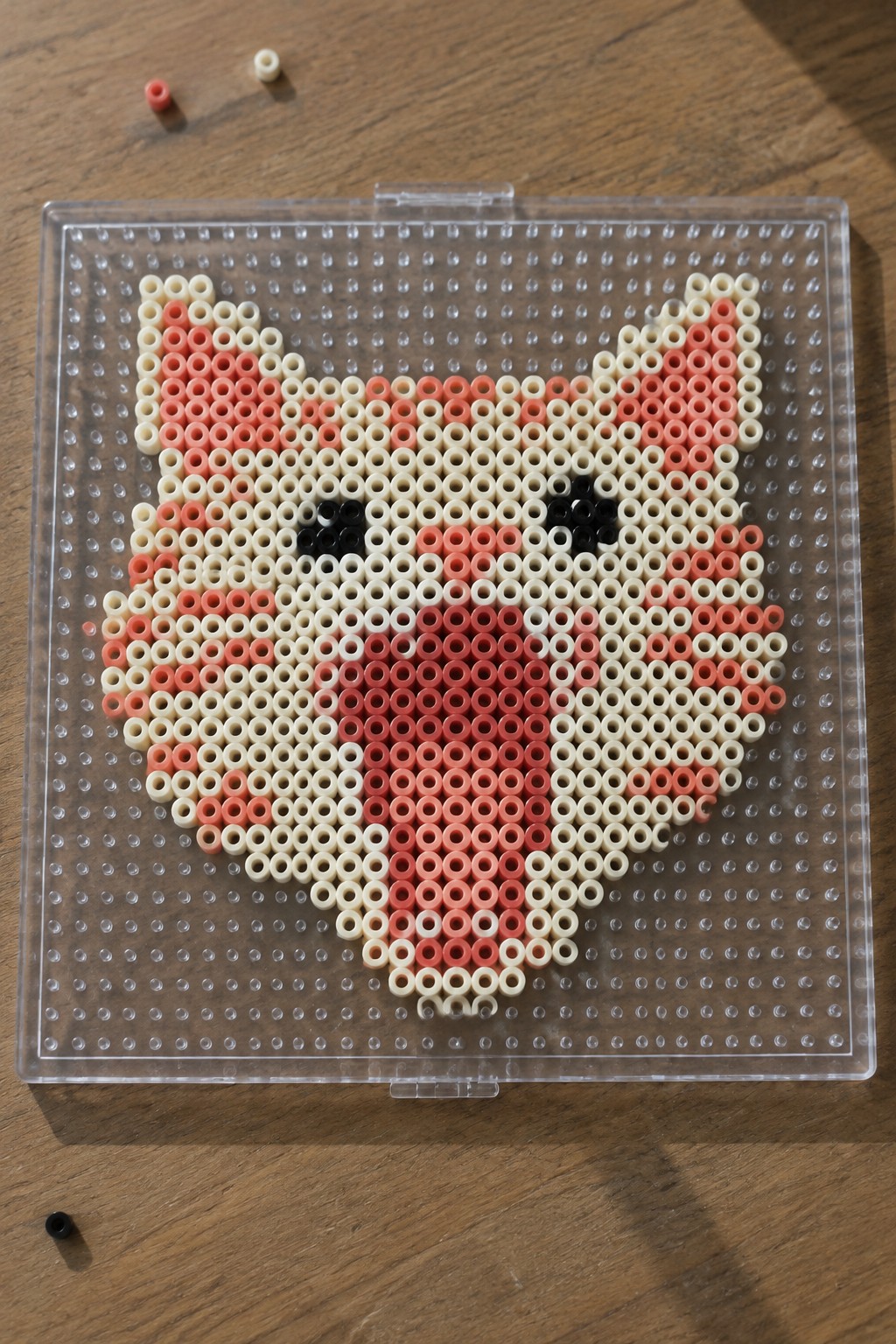 Pink red white Perler beads forming cat face on clear pegboard over wood