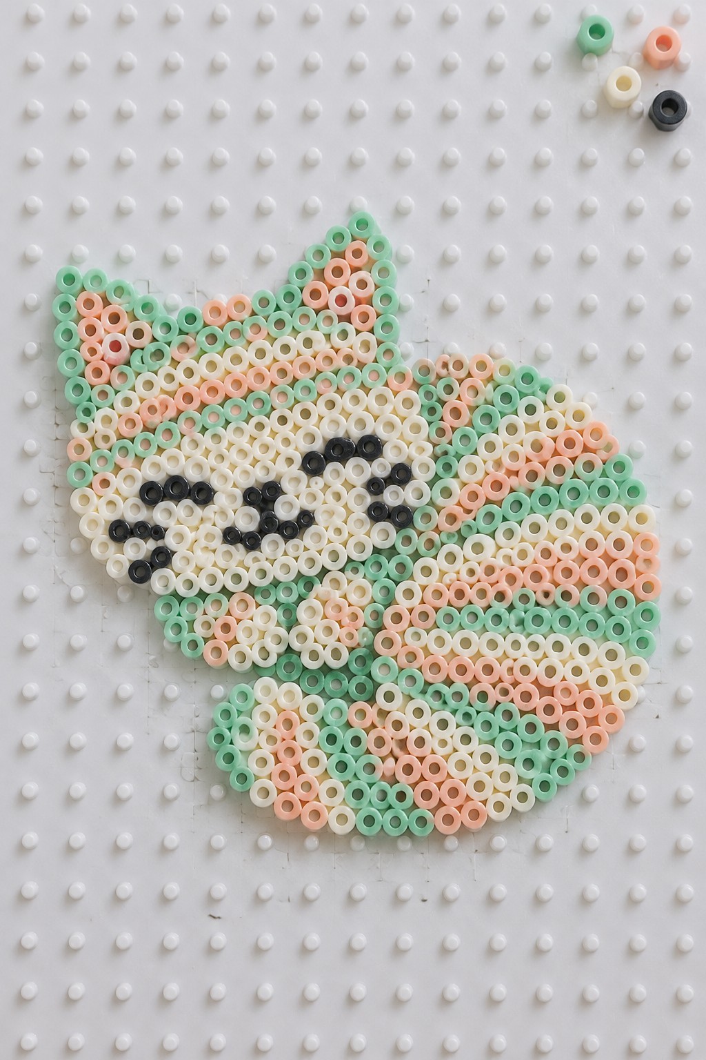 Pastel perler bead cat face in mint green, peach, and white on pegboard
