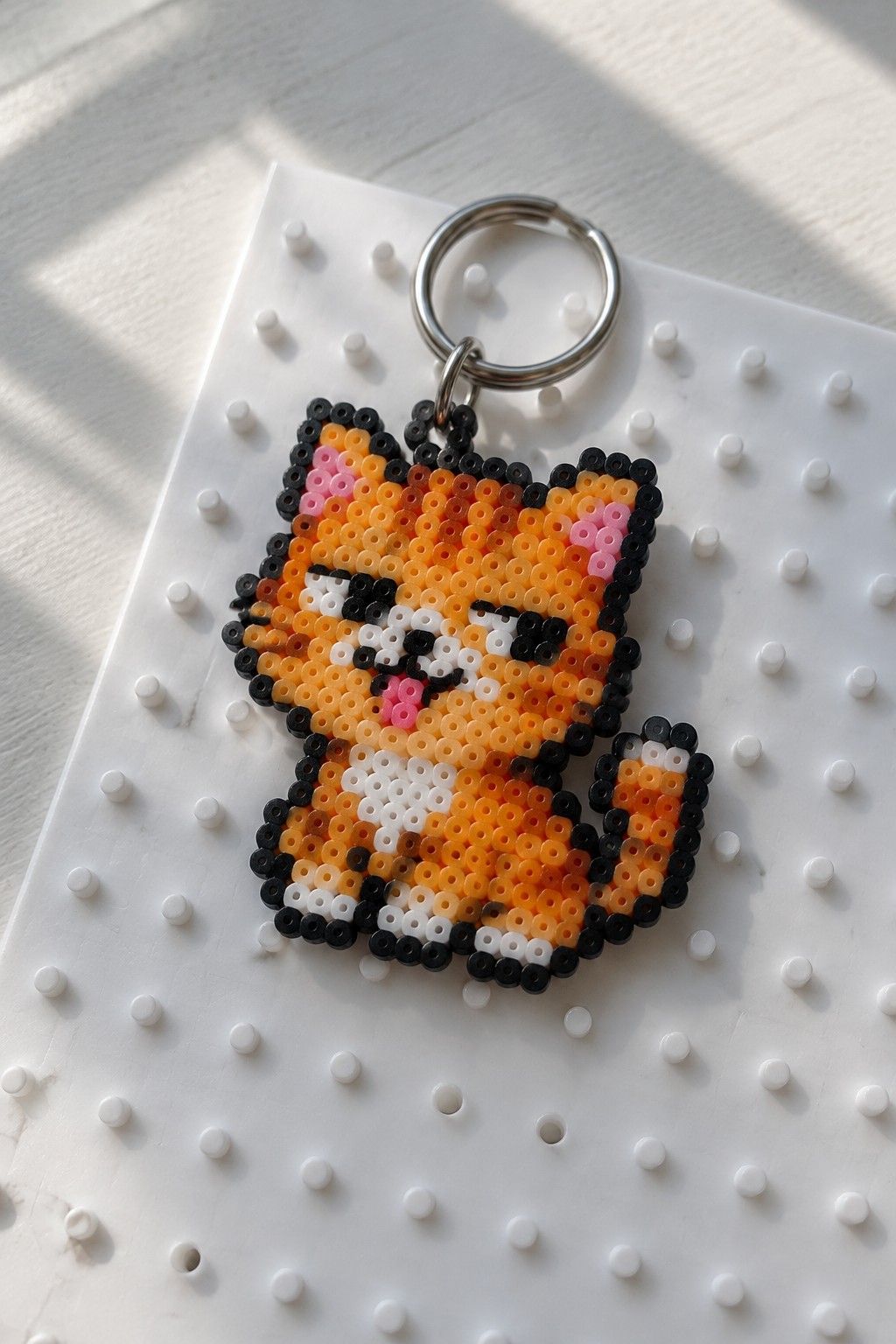 Orange perler bead cat keychain with pink cheeks on white pegboard