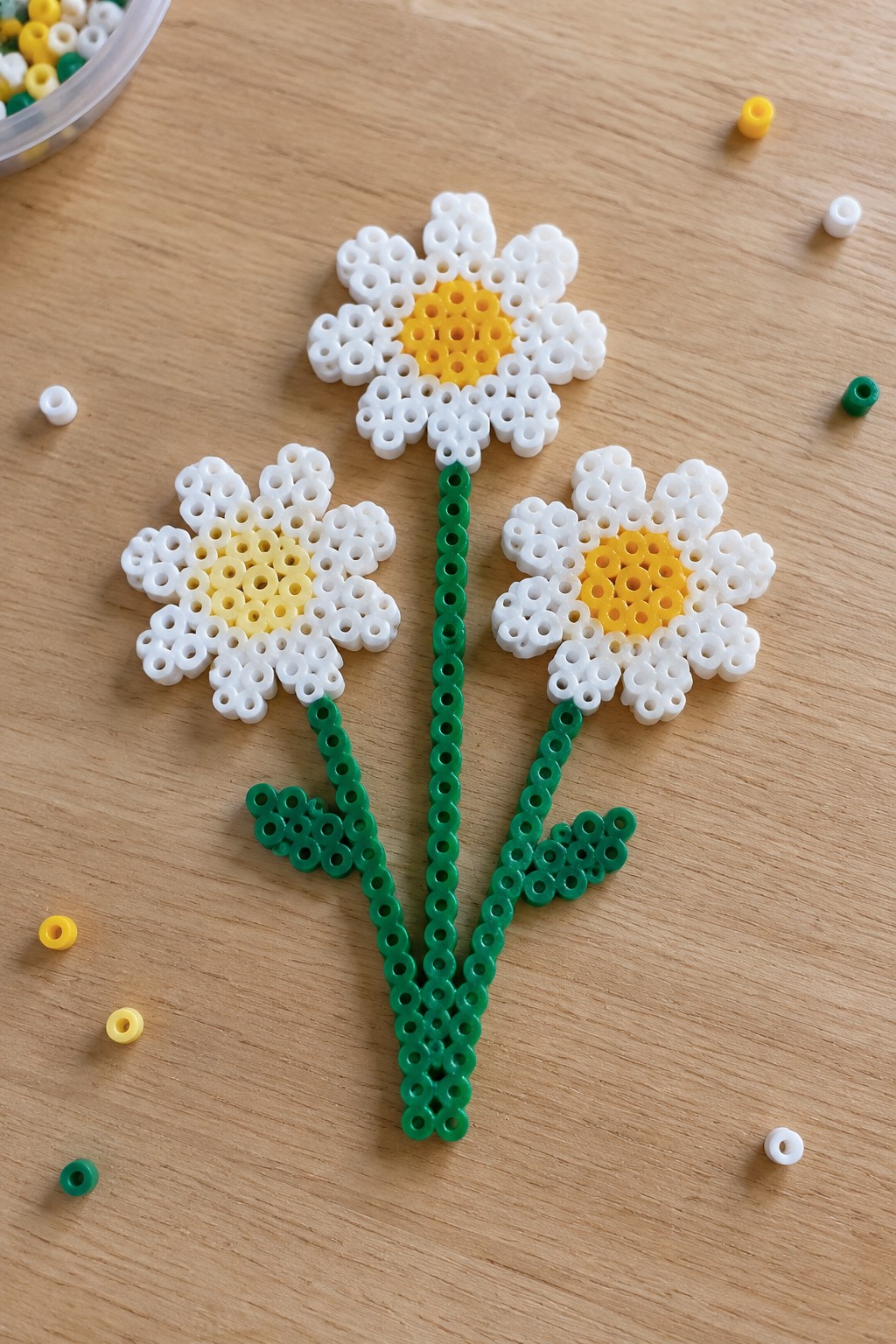 Three daisy Perler bead flowers with white petals, yellow centers, and green stems on a pegboard.