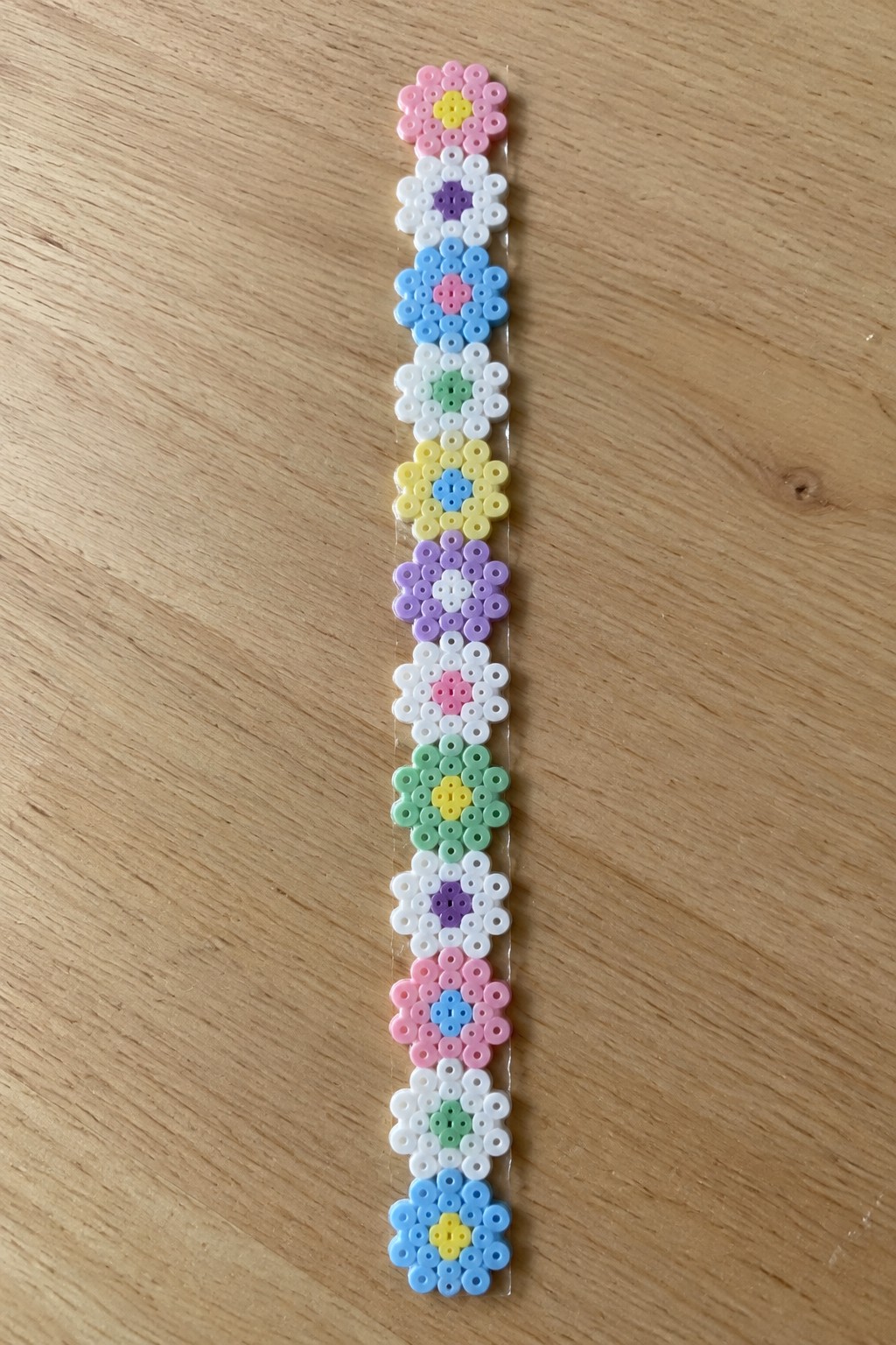 A vertical strip of fused Perler beads displaying multiple daisy flowers in different color combinations.