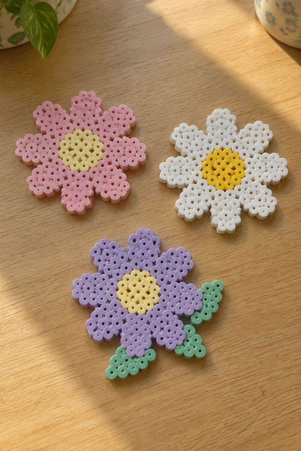 Three perler bead flowers in pink, white, and purple with yellow centers on wood.