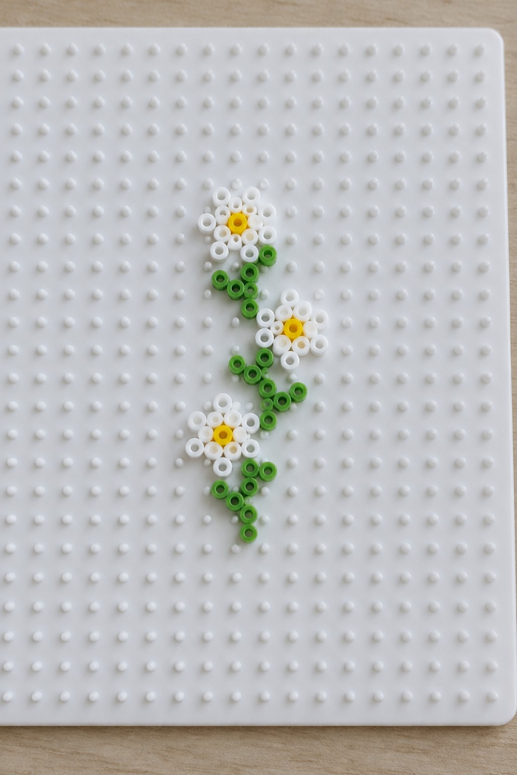 Three beaded daisies with white petals, yellow centers, and green stems on pegboard.