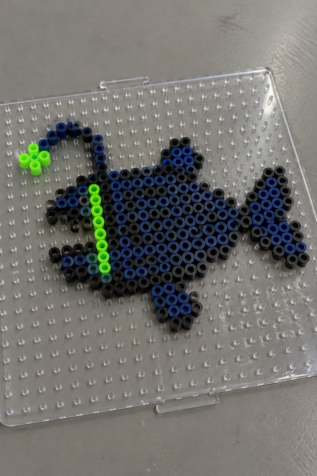 Perler beads in blue, black, and neon green forming shapes on a clear pegboard