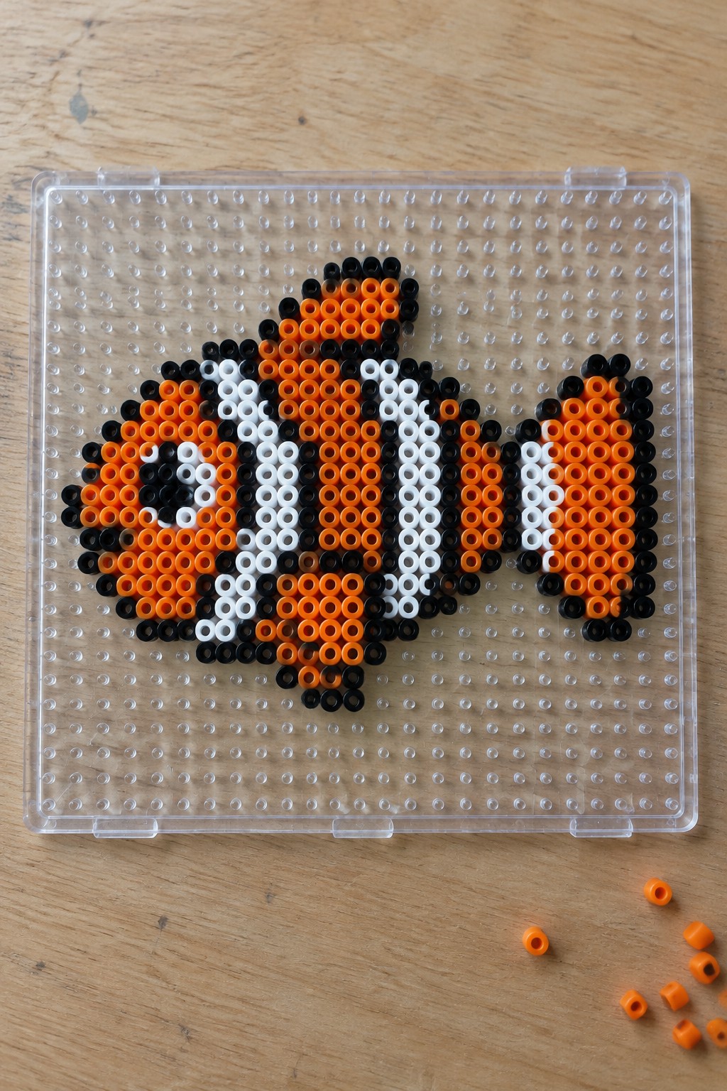 A clownfish made from orange, white, and black Perler beads arranged on a clear pegboard.