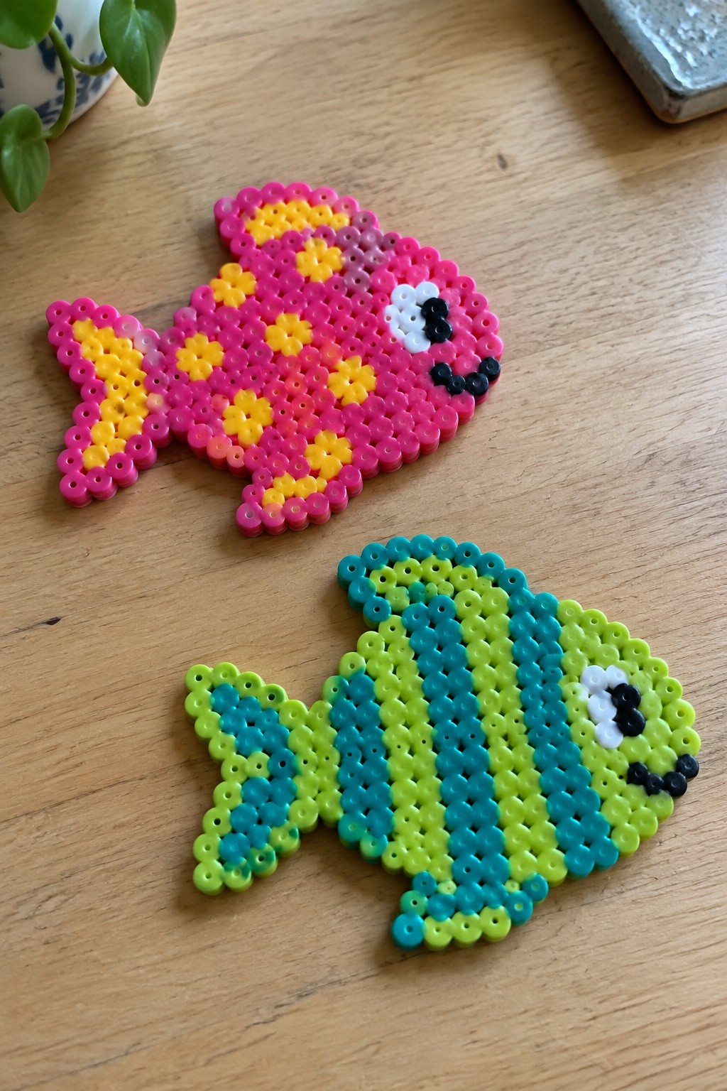 Two perler bead fish crafts, pink-yellow and green-blue, on wooden table