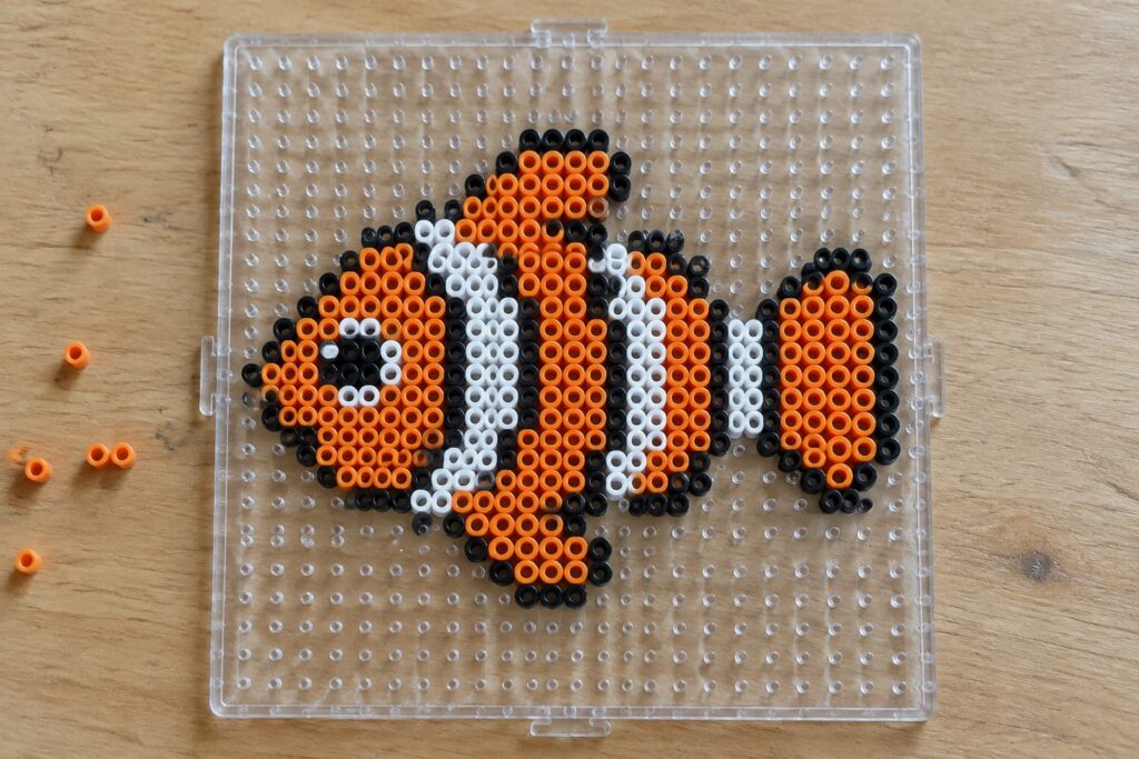 A clownfish made from orange, white, and black Perler beads arranged on a clear pegboard.