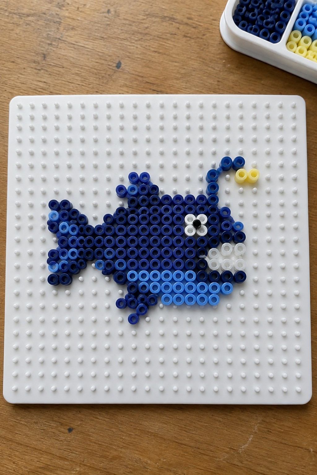 Blue perler beads forming a fish on white pegboard with yellow accents nearby.