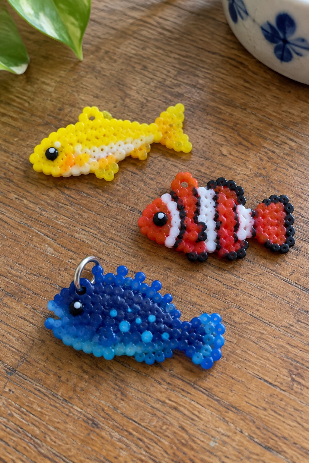 Three perler bead fish: yellow, clownfish-striped, and blue gradient on wood.