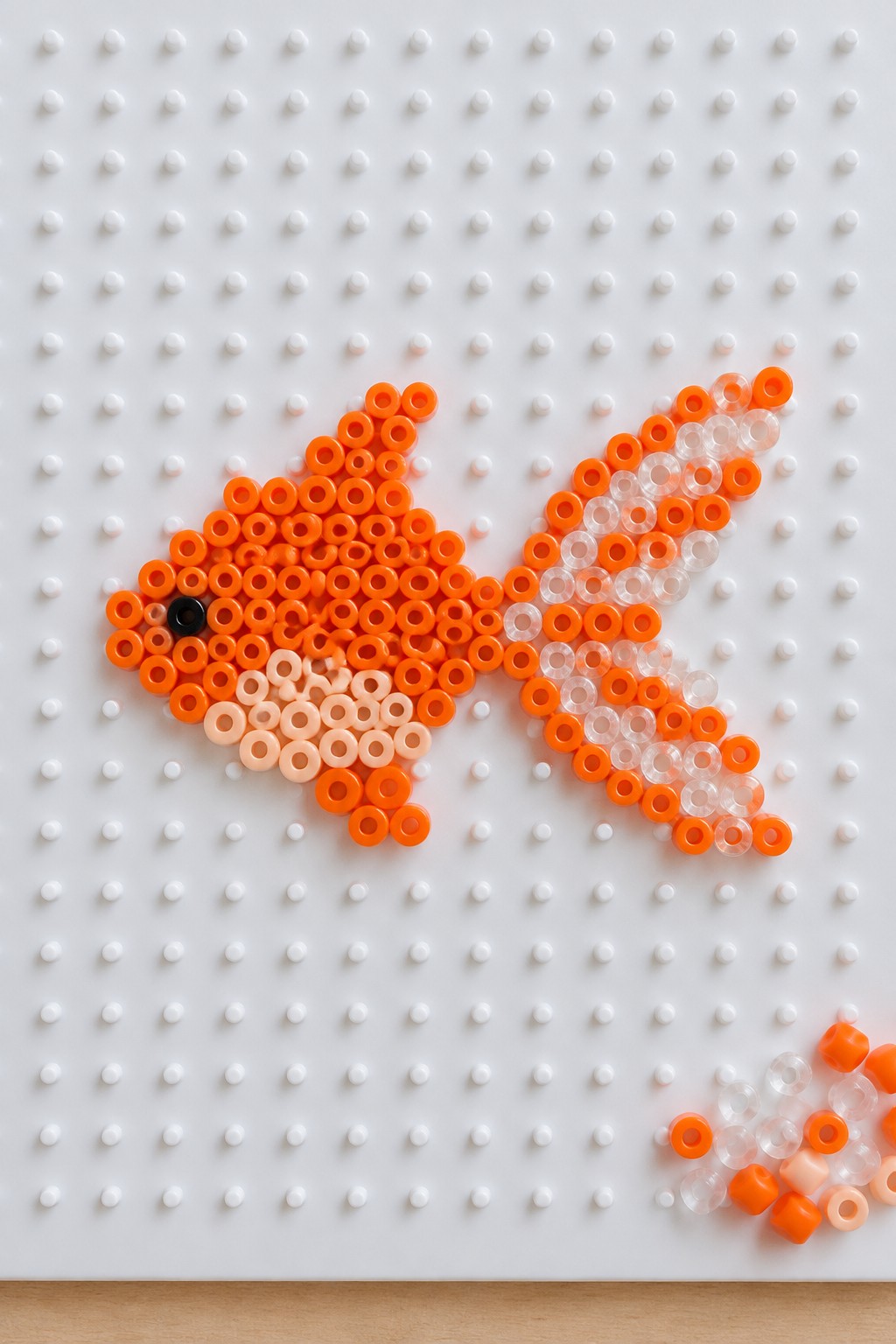 Orange perler bead goldfish with black eye on white pegboard and loose beads