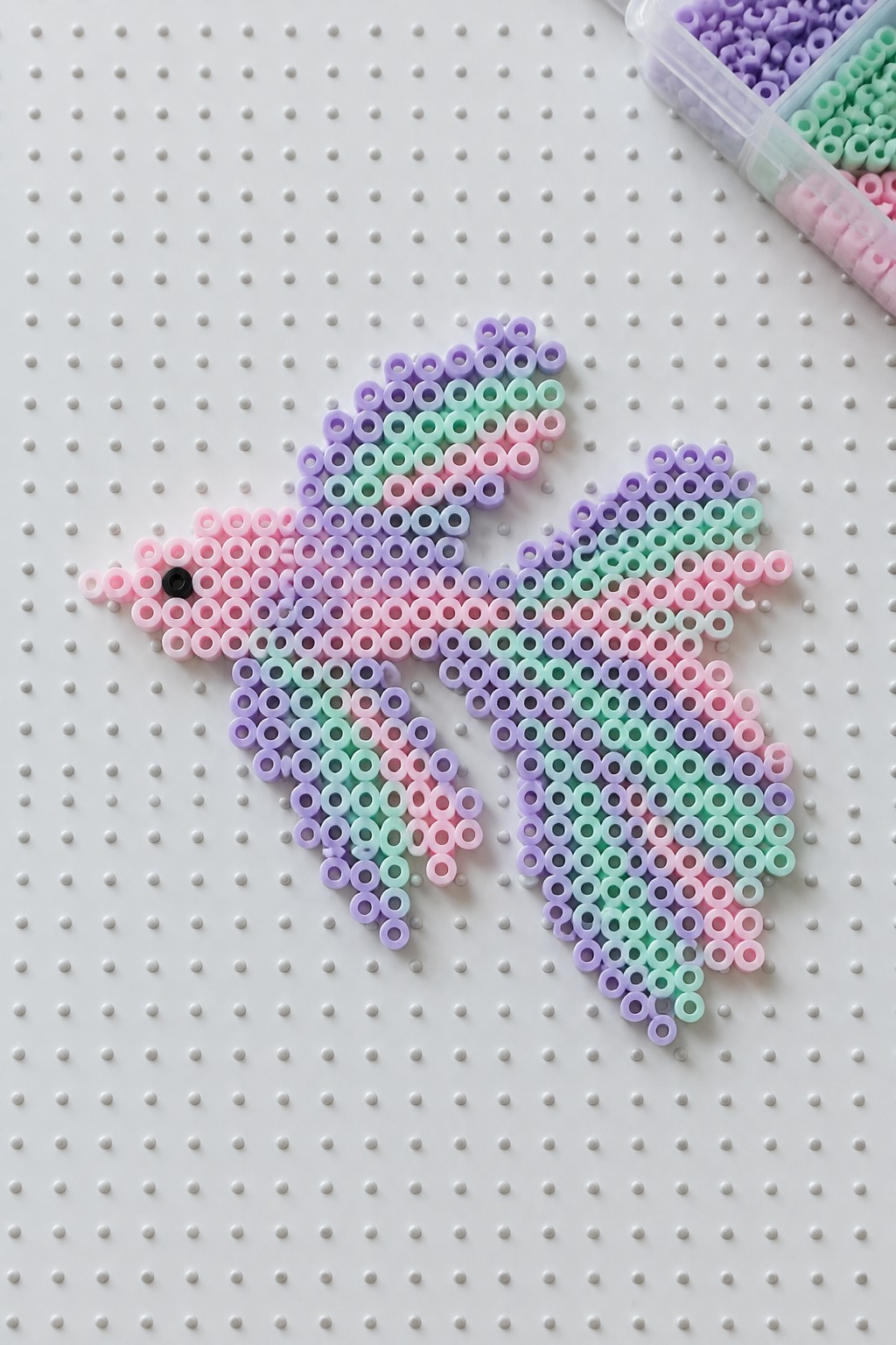 Pastel perler beads form a bird on white pegboard near open bead organizer