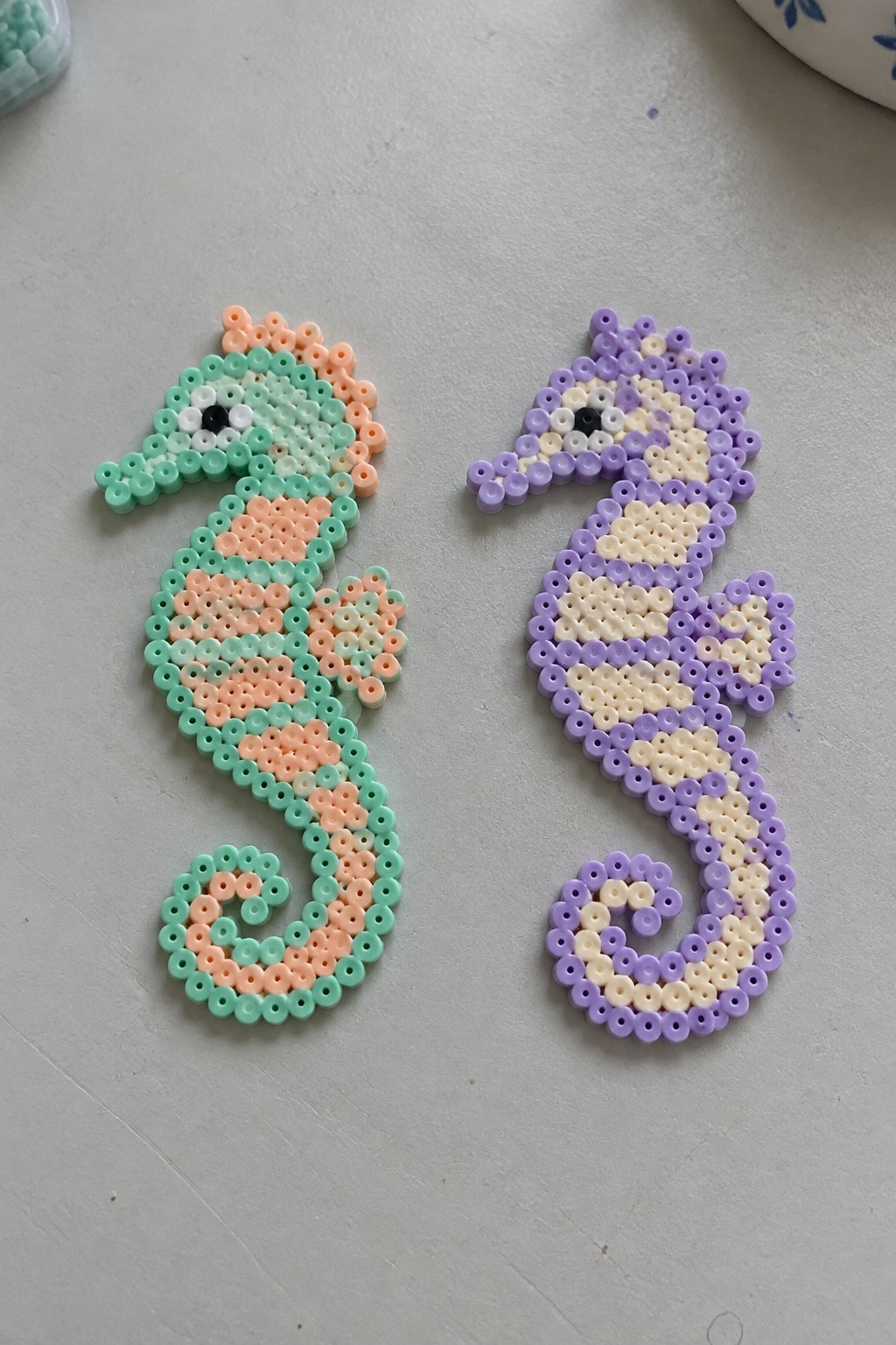 Two seahorse Perler bead patterns, one in mint green and peach with a face, the other in purple and cream with a face.