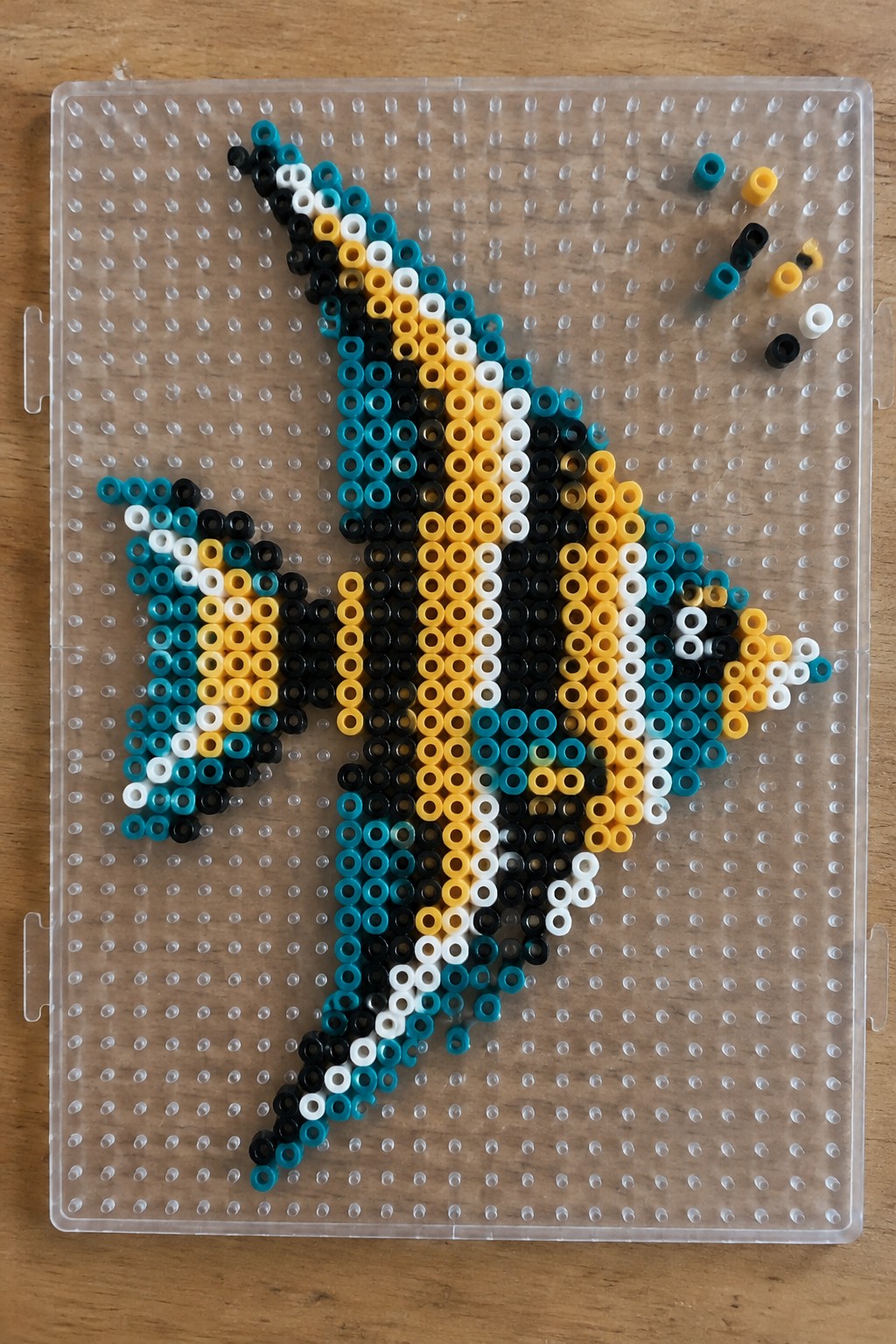 Yellow, black, white, and teal perler beads forming a fish on clear pegboard