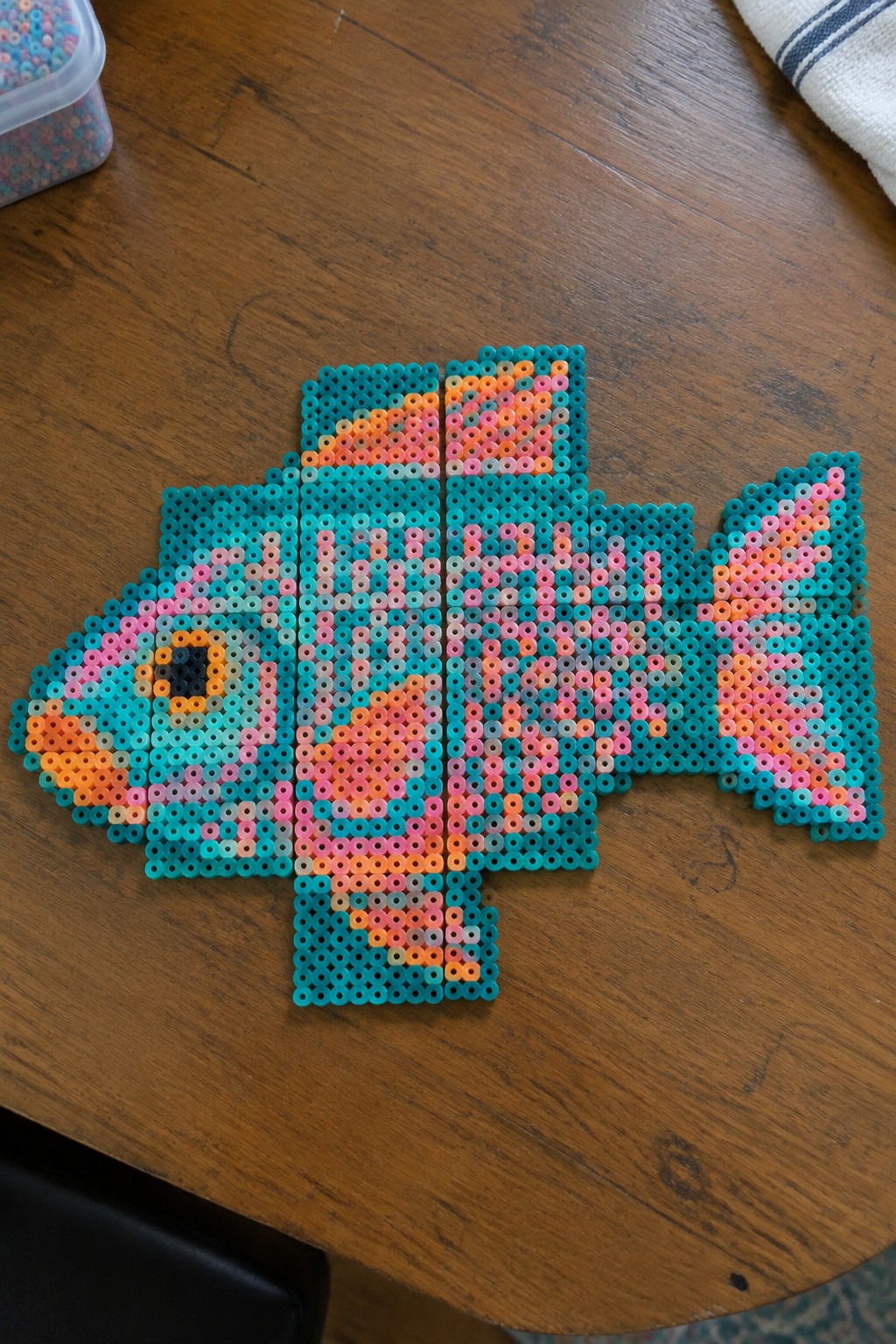 Colorful perler bead fish design on wooden table with bead container nearby