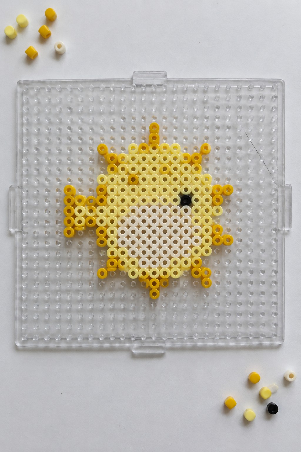 Yellow perler bead chick with black eye on clear pegboard amid loose beads