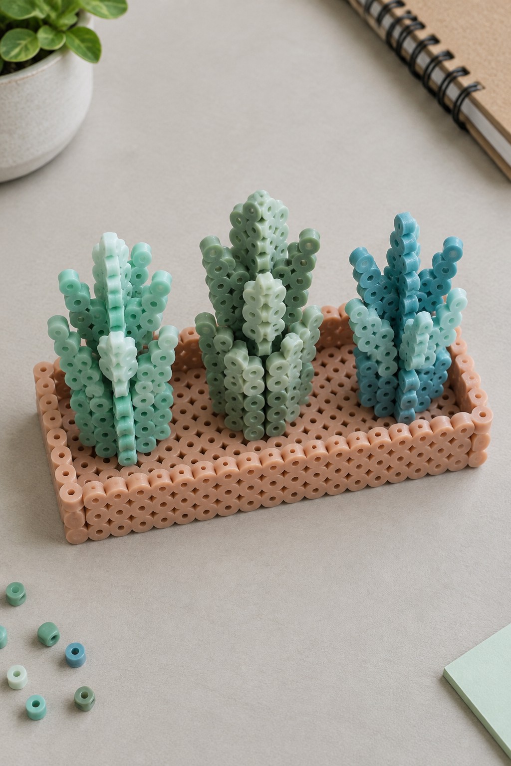 Three perler bead cacti in green and blue on a peach tray with loose beads
