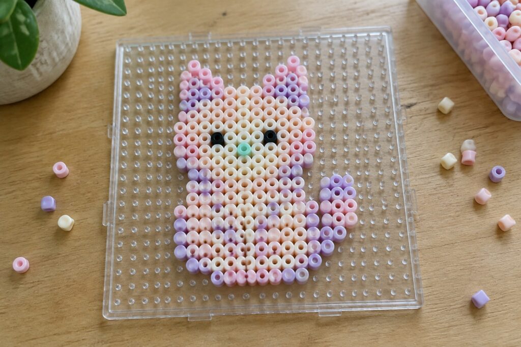 18 Cute Kawaii Perler Bead Ideas in Soft Pastel Colors Pastel pink and purple perler bead cat on clear pegboard over wood.
