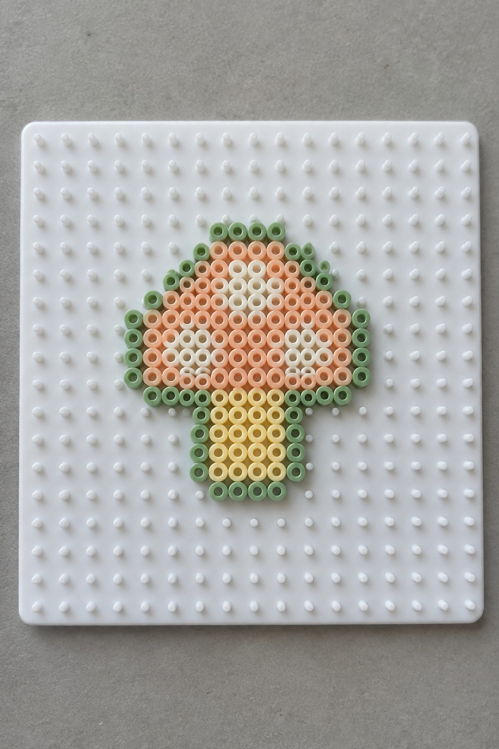 Perler bead mushroom with green-orange cap and yellow stem on white pegboard