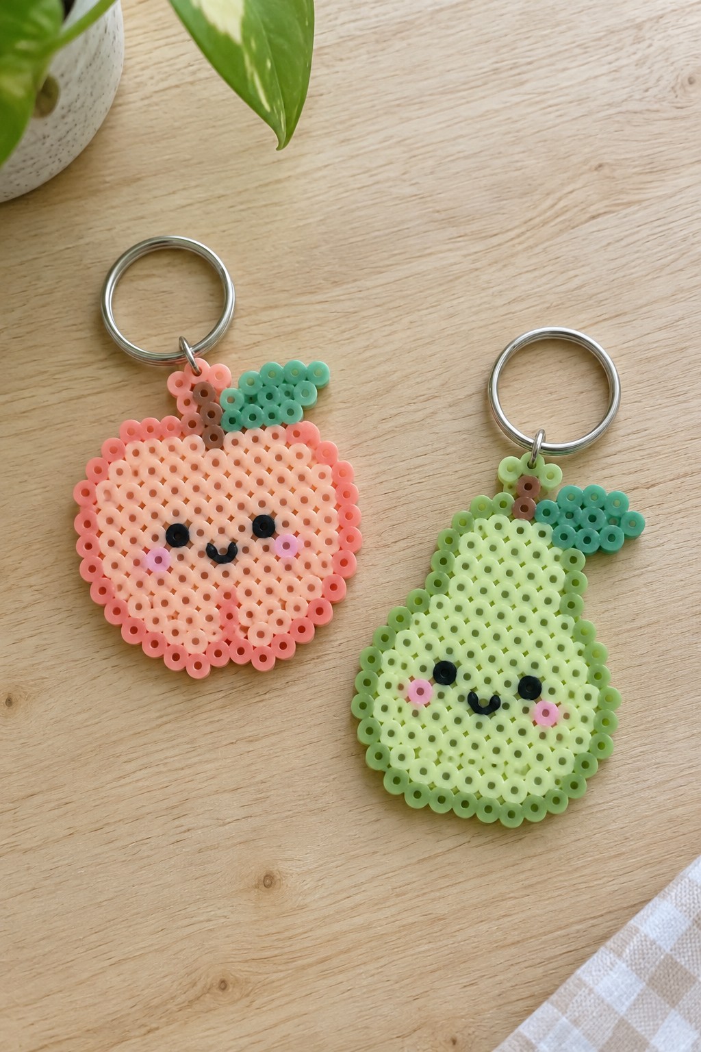 Two fused Perler bead keychains in the shape of a peach and a pear with kawaii faces, made in soft pastel pink, coral, green, and teal beads.