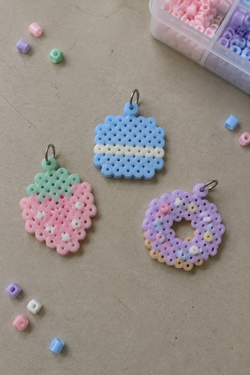 Three pastel perler bead keychains—strawberry, striped square, sprinkle donut—with scattered beads