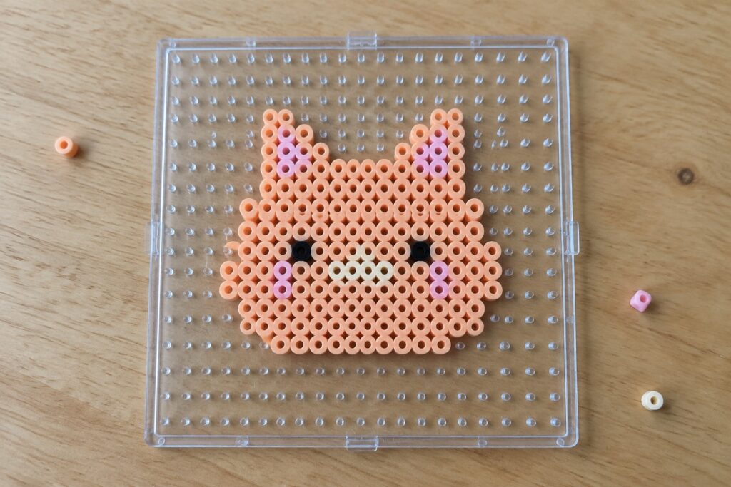 18 Cute Perler Bead Ideas Featuring Kawaii Characters Peach perler bead cat face with pink cheeks on clear pegboard