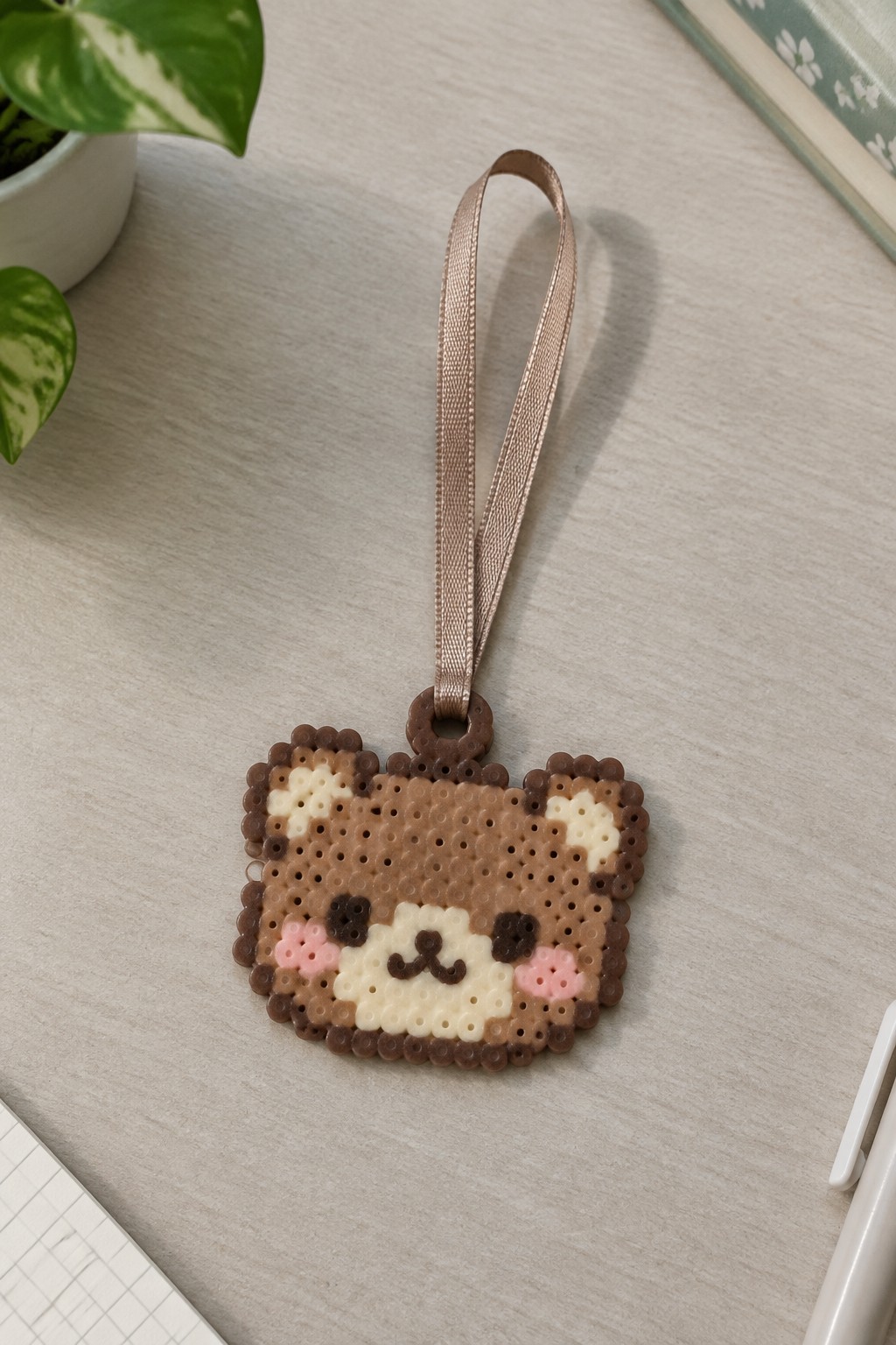 Perler bead bear face with pink cheeks and beige ribbon on wooden table.