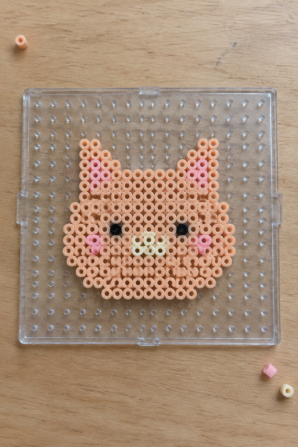 Peach perler bead cat face with pink cheeks on clear pegboard