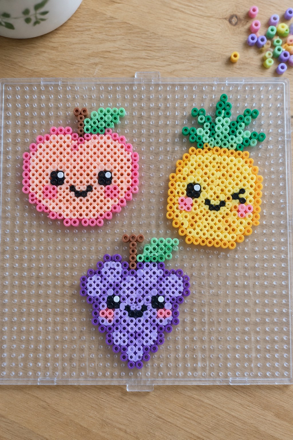 Three kawaii fruit Perler bead designs on a pegboard showing a peach, pineapple, and grapes with simple faces.
