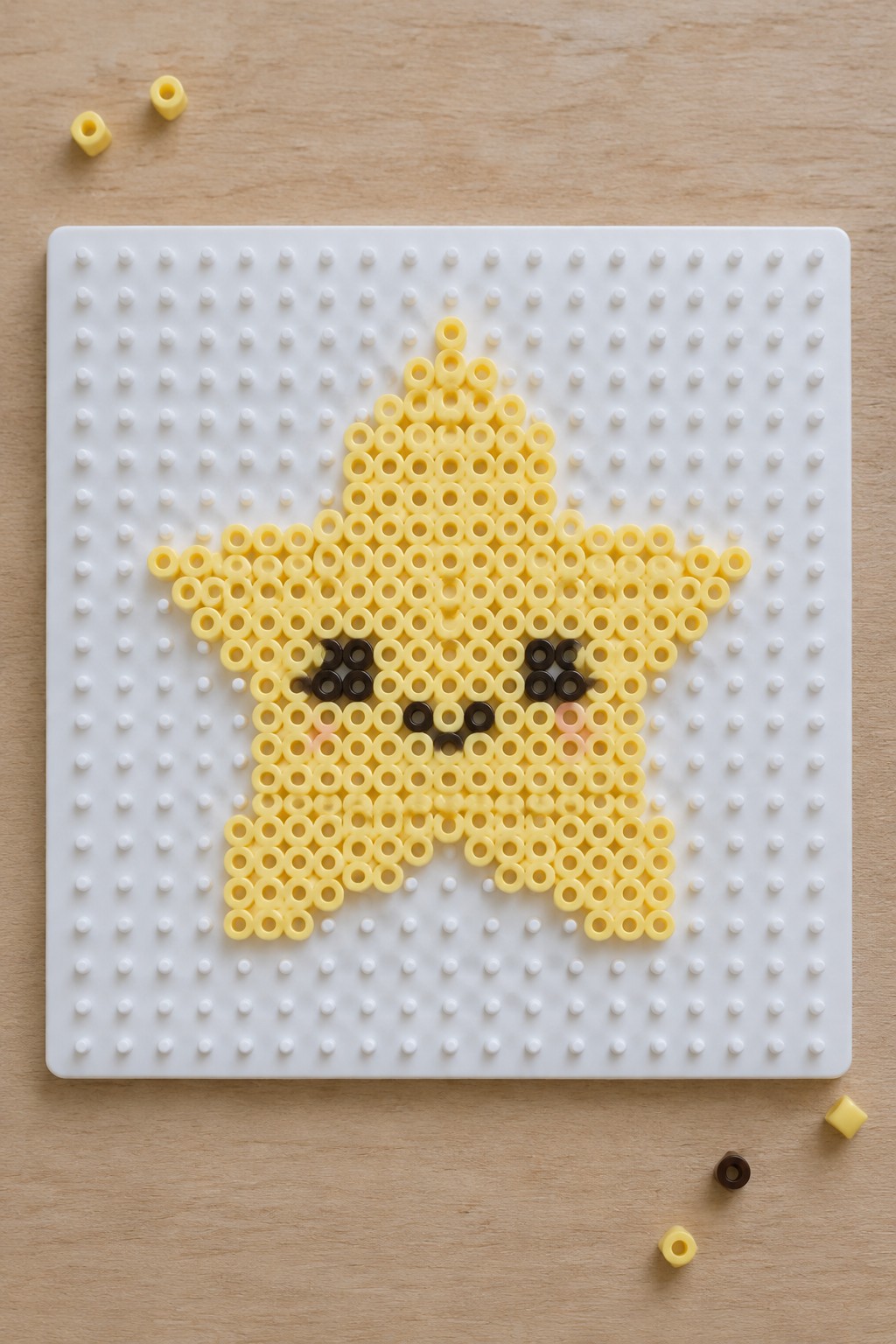 Yellow Perler bead star with black eyes, black mouth, and pink cheeks arranged on a white pegboard.