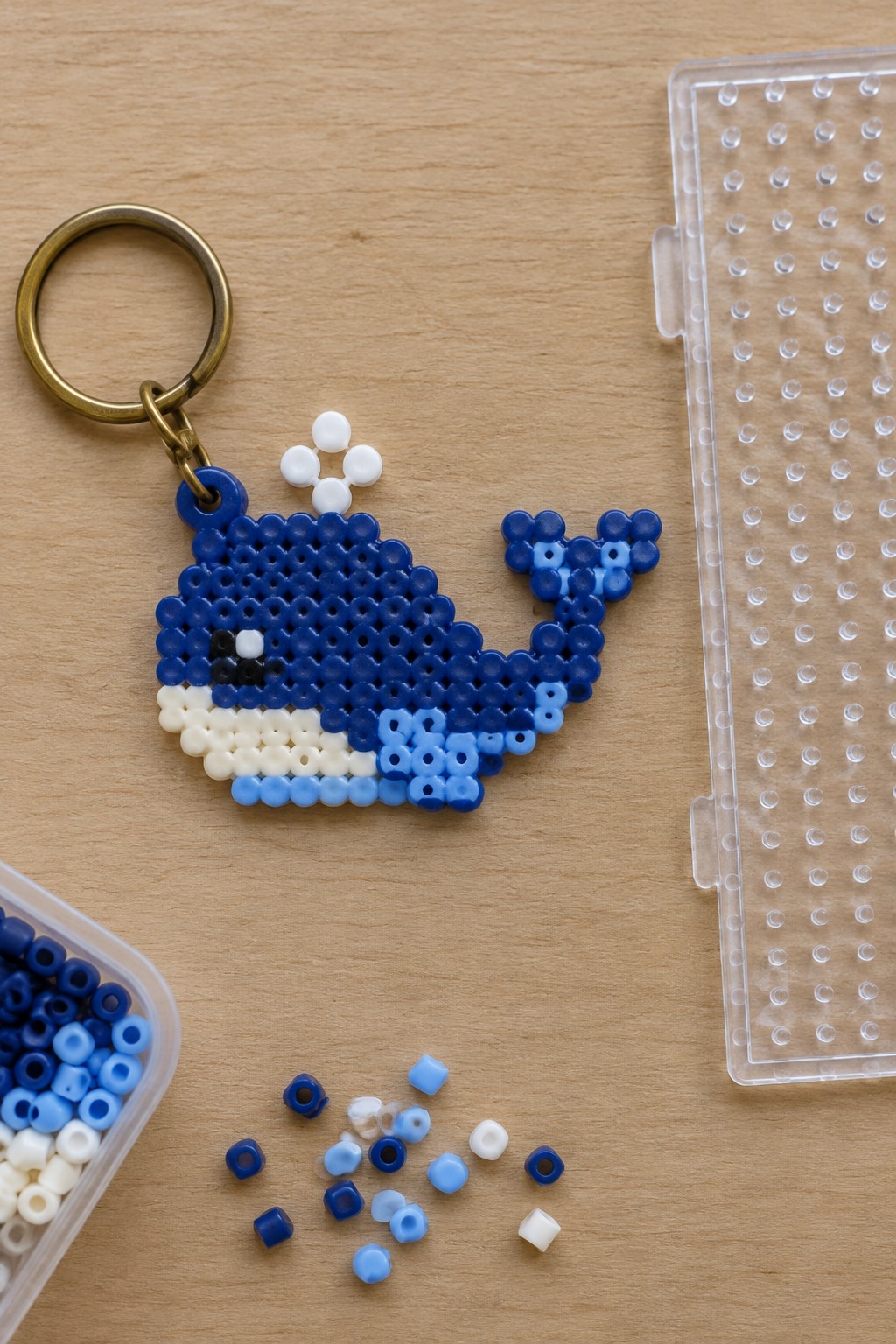 Blue perler bead whale keychain beside clear pegboard and scattered beads on wood