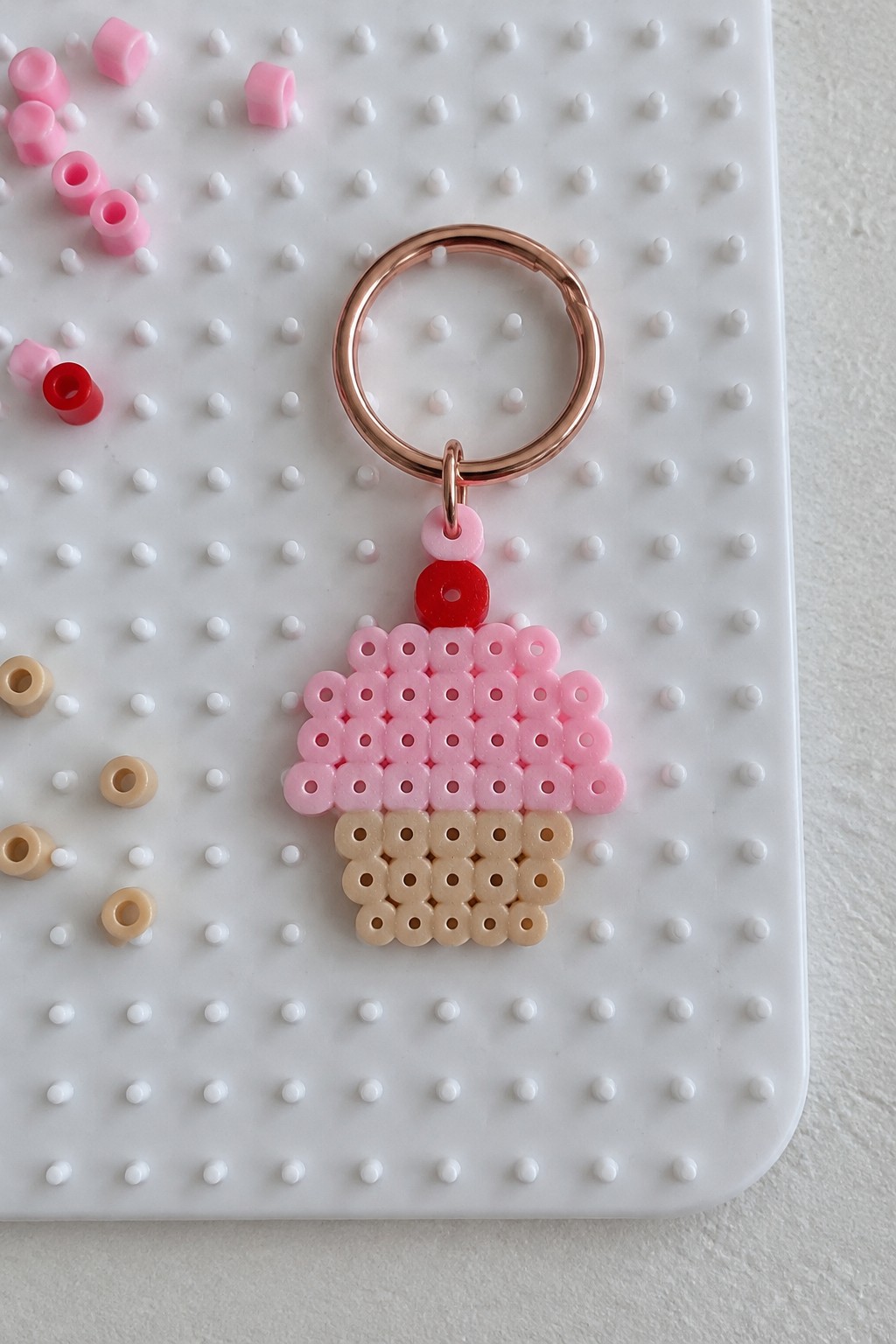 Pink and beige cupcake Perler bead keychain on a pegboard with a rose gold ring.