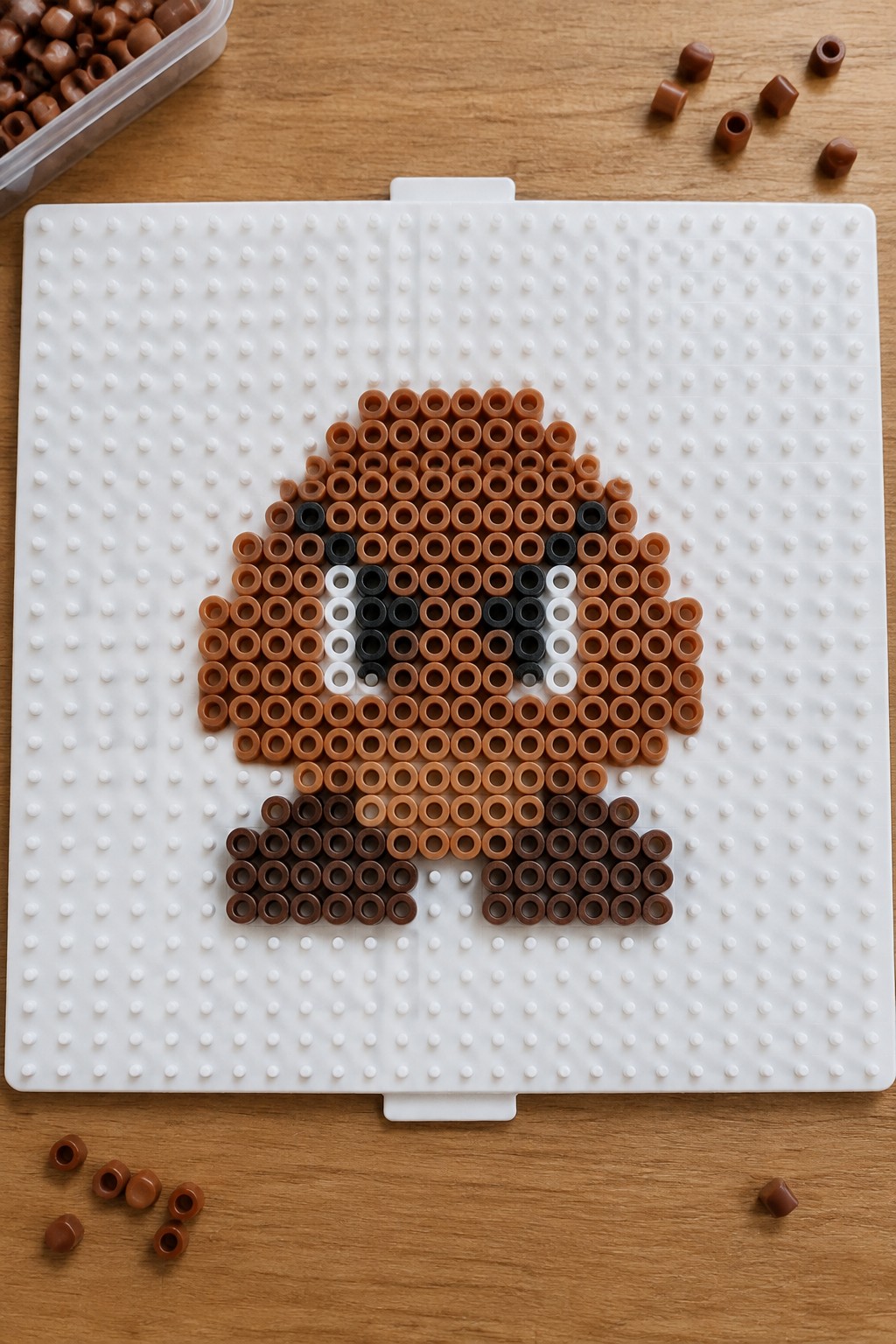 Perler beads forming a brown cartoon face with black eyes on white pegboard.